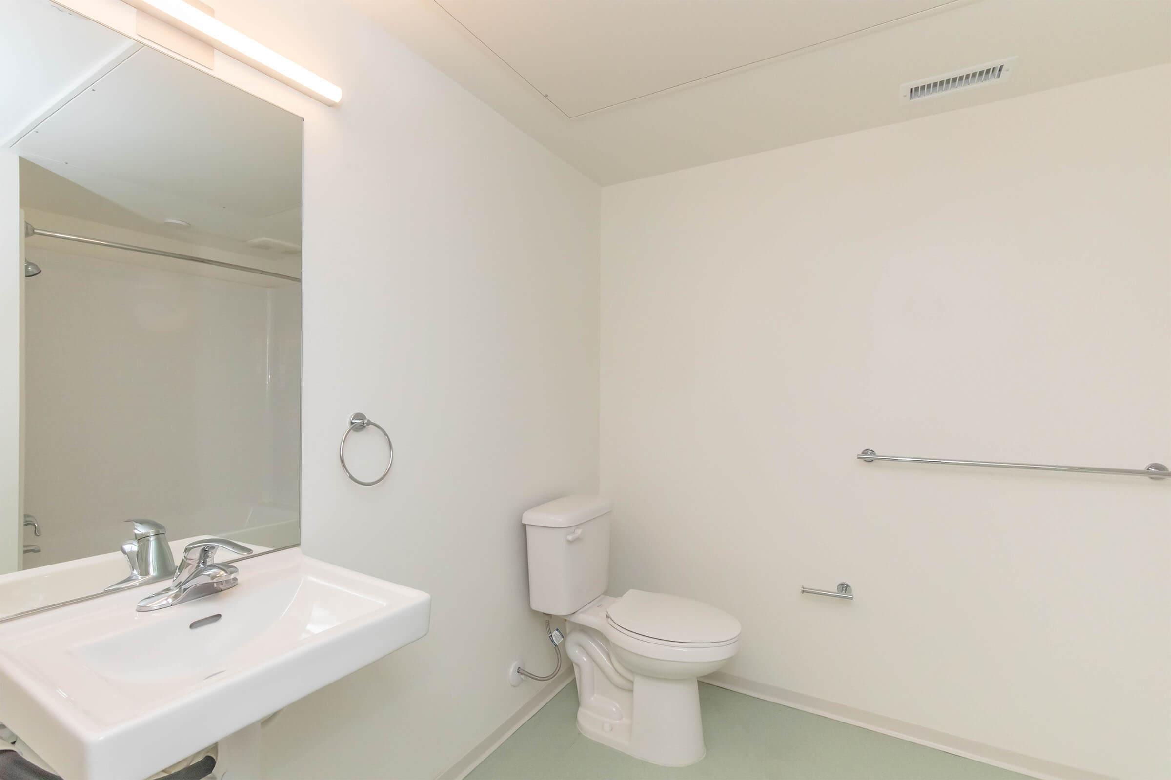A clean and minimalistic bathroom featuring a white sink with a modern faucet, a toilet, and a shower with a clear glass door. The walls are painted white, and the floor is a light green color. A large mirror is mounted above the sink, providing a bright, uncluttered appearance.