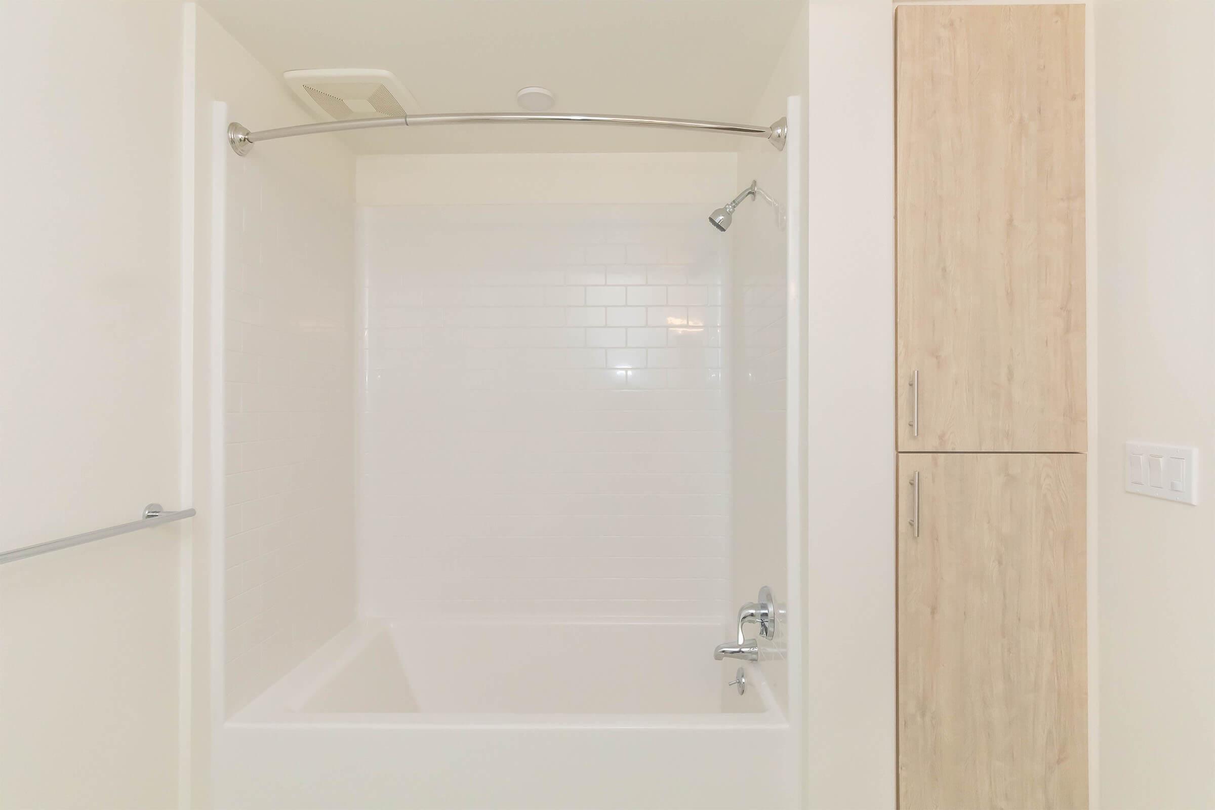 A clean and modern bathroom featuring a white tiled shower area with a curved shower rod, a handheld showerhead, and a bathtub. To the right, there is a light wood storage cabinet. The overall aesthetic is minimalistic and bright.