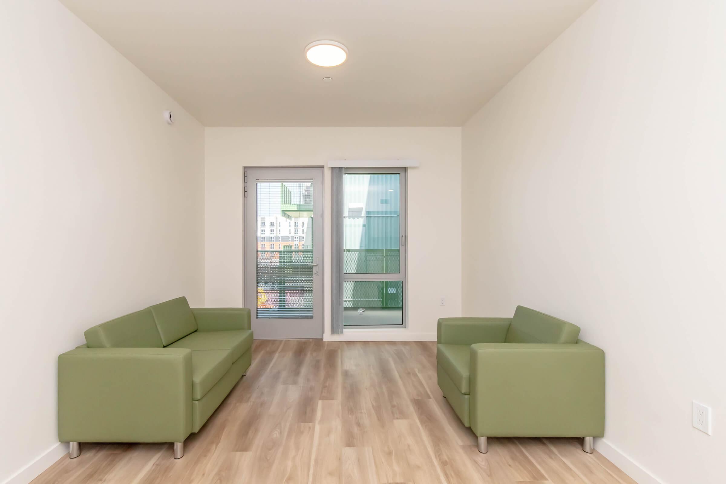A minimalist living space featuring two green sofas facing each other. Large windows allow natural light, with a view of the outdoors. The floor is made of light wood, and the walls are painted in a soft, neutral tone, creating a modern and airy atmosphere.