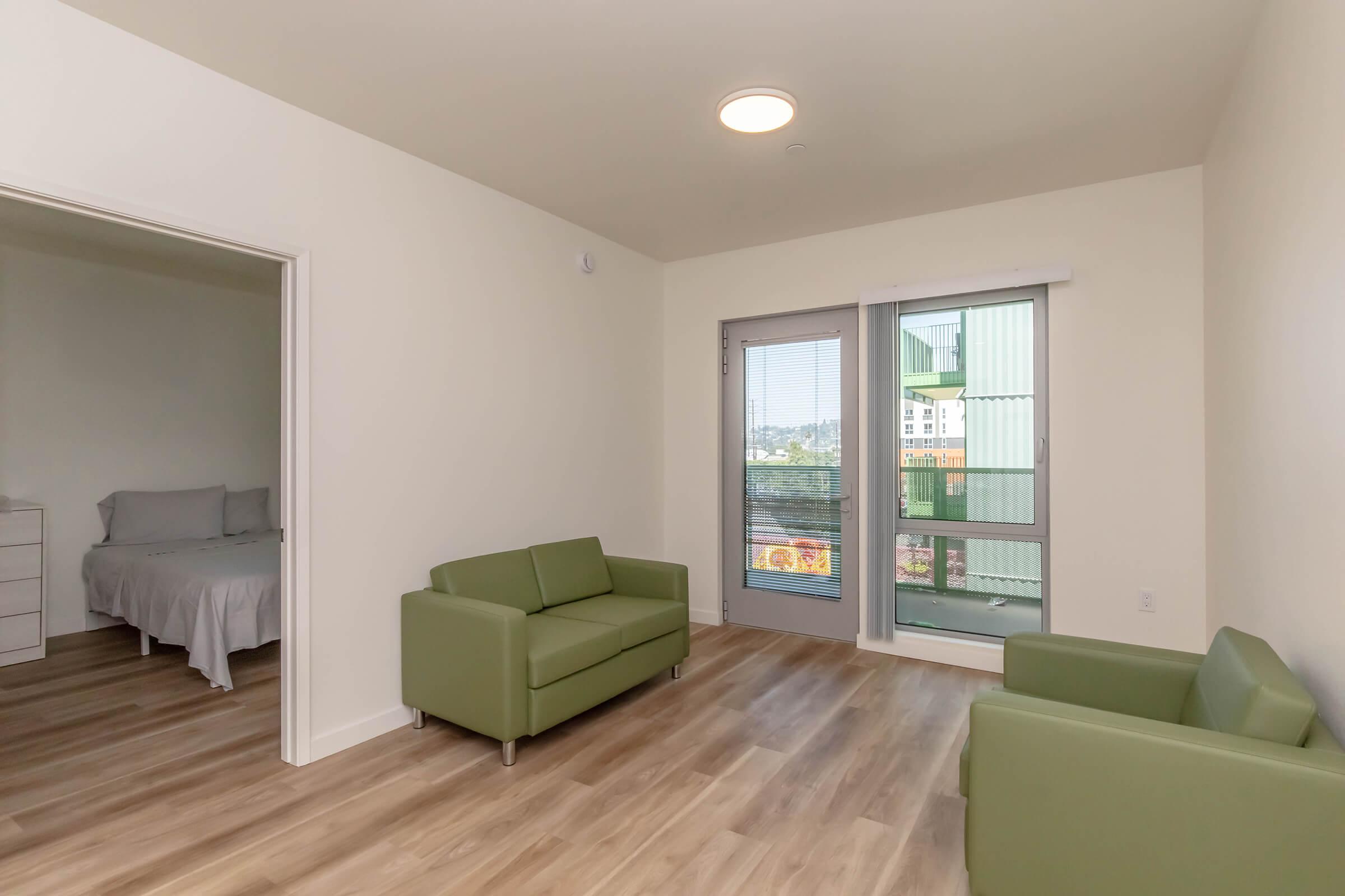 A modern living room featuring two green sofas, light-colored walls, and a large window with a view. A doorway leads to a bedroom, and the floor is made of light wood. Natural light brightens the space, creating a cozy atmosphere.