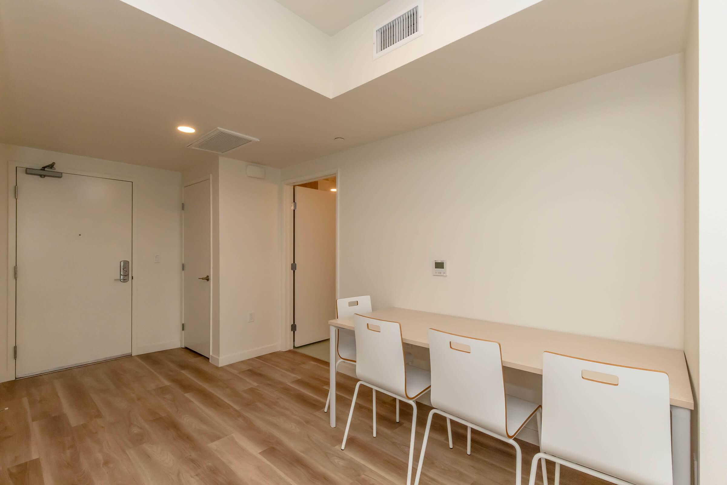 An empty room featuring light-colored walls and wooden flooring. There is a simple table against one wall with three white chairs. A door is visible on the left, and another door leads to a different space. Natural light enters, creating a bright and minimalist atmosphere.