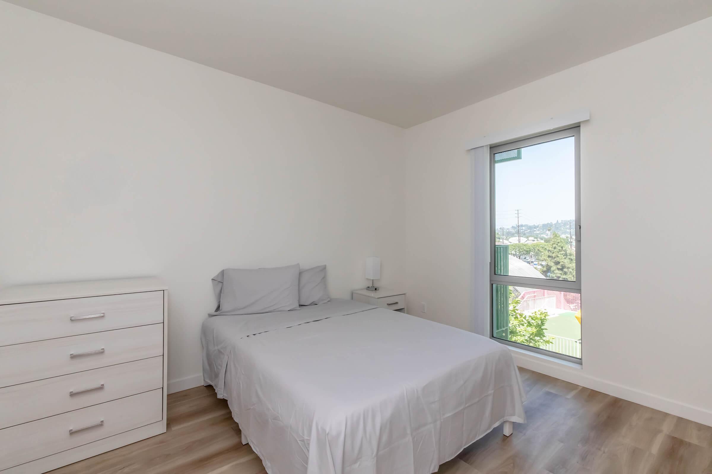 A minimalist bedroom featuring a light-colored bed with a gray blanket, a nightstand with a lamp, and a chest of drawers. A large window allows natural light, showing a view of greenery outside. The walls are painted white, contributing to a bright and airy atmosphere.