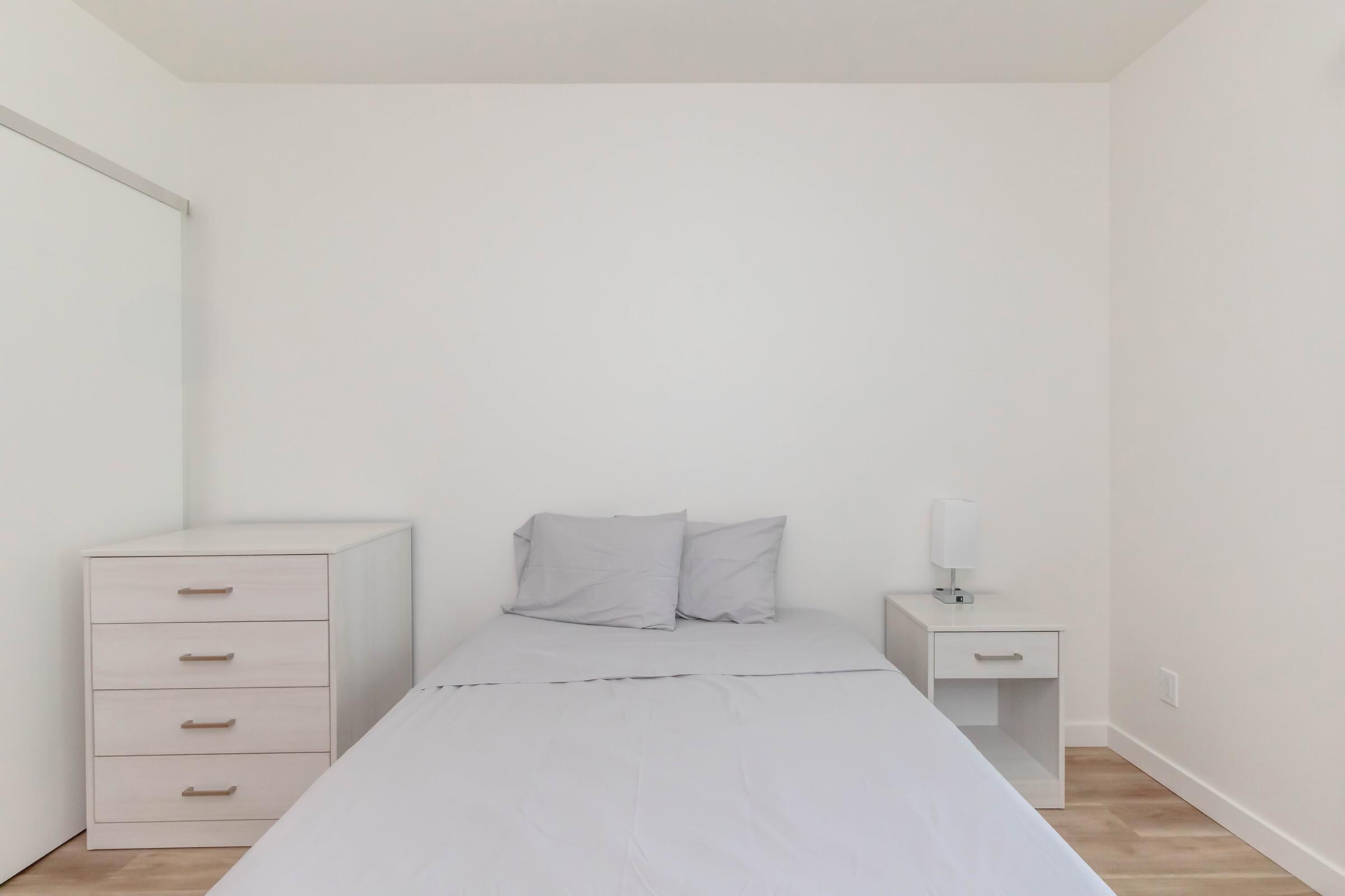 A minimalist bedroom featuring a light-colored bed with gray bedding and two pillows, accompanied by a small bedside table with a lamp. A simple chest of drawers is positioned to the left, against a white wall. The floor is light wood, contributing to the airy atmosphere of the room.