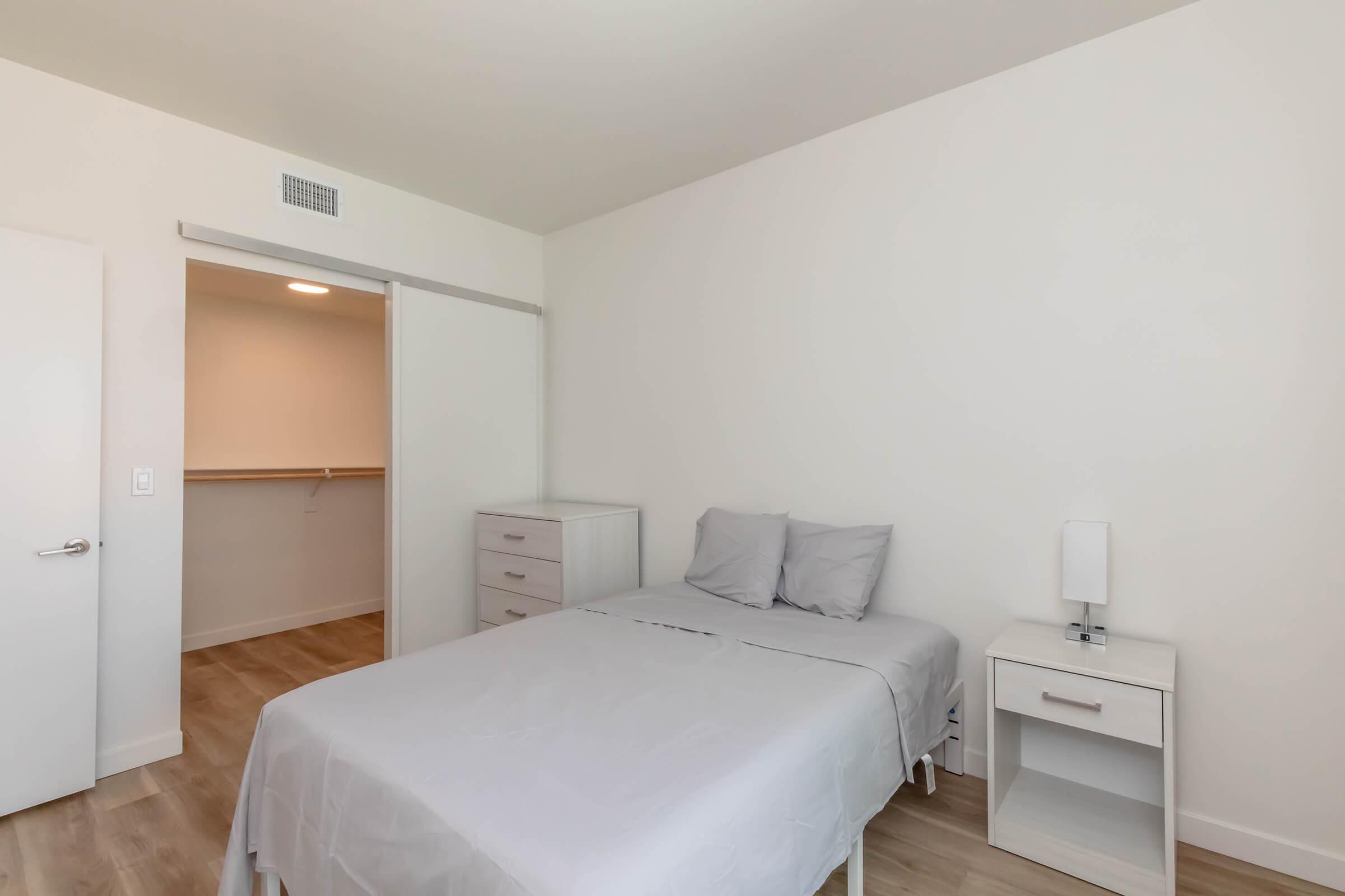 A minimalist bedroom featuring a bed with gray bedding, a nightstand with a lamp, and an open closet area. The walls are white, and the floor is light-colored wood, creating a spacious and clean aesthetic.
