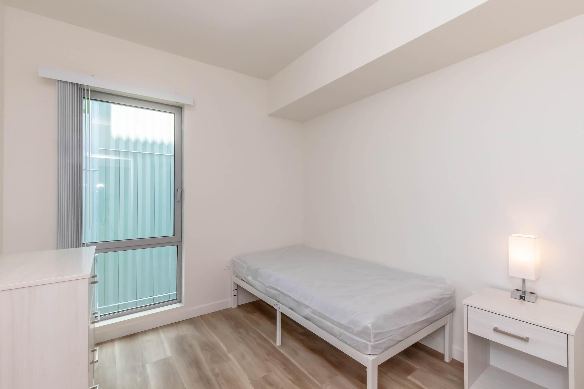 A simple, unfurnished bedroom featuring a single bed with a light gray mattress, a nightstand with a lamp, and a large window allowing natural light. The walls are painted white, and the flooring is light wood.