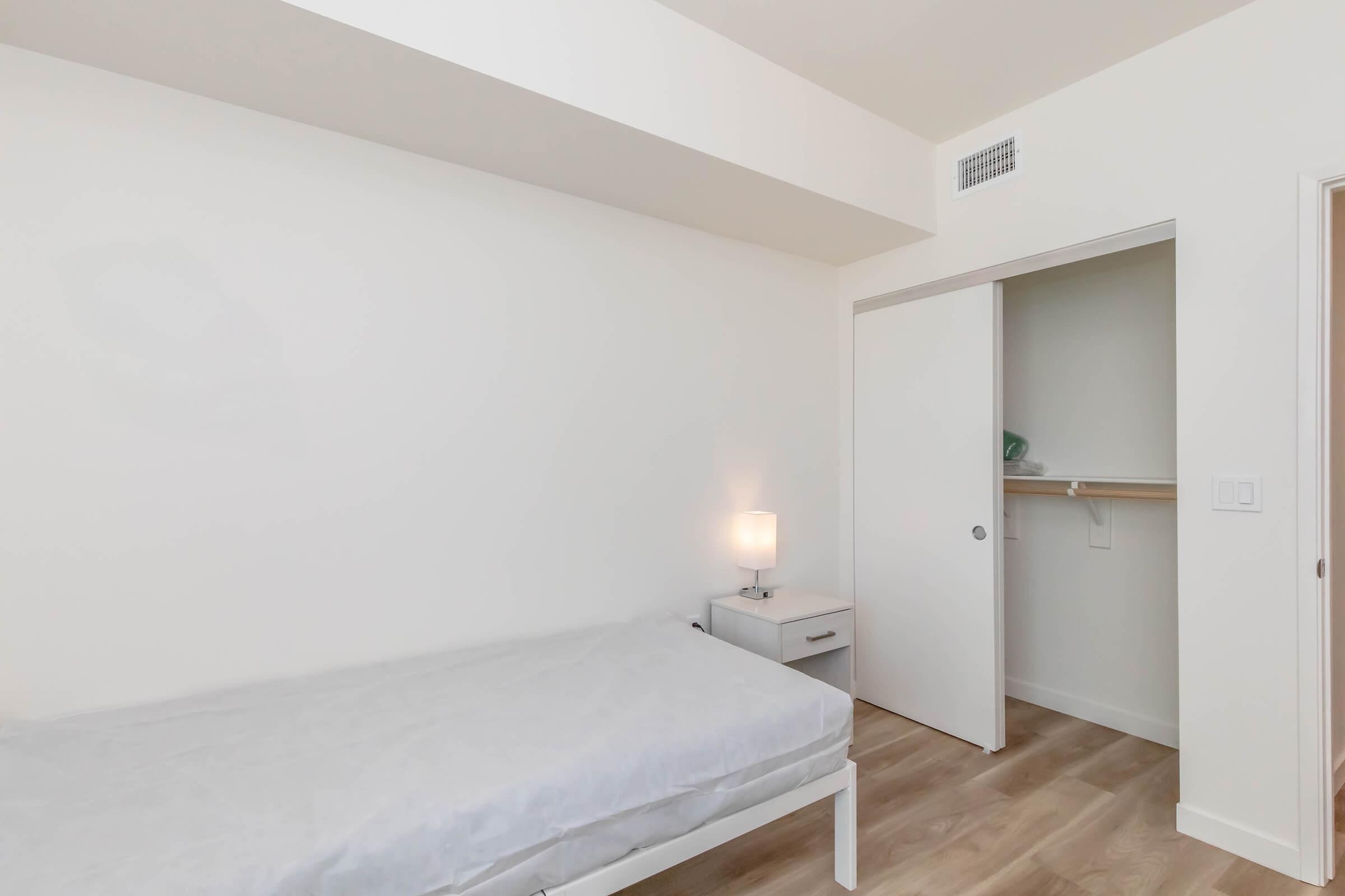 A minimalistic bedroom featuring a single bed with a light-colored mattress, a nightstand with a small lamp, and a closet with sliding doors. The walls are painted white, and there is light wood flooring, creating a bright and open atmosphere.