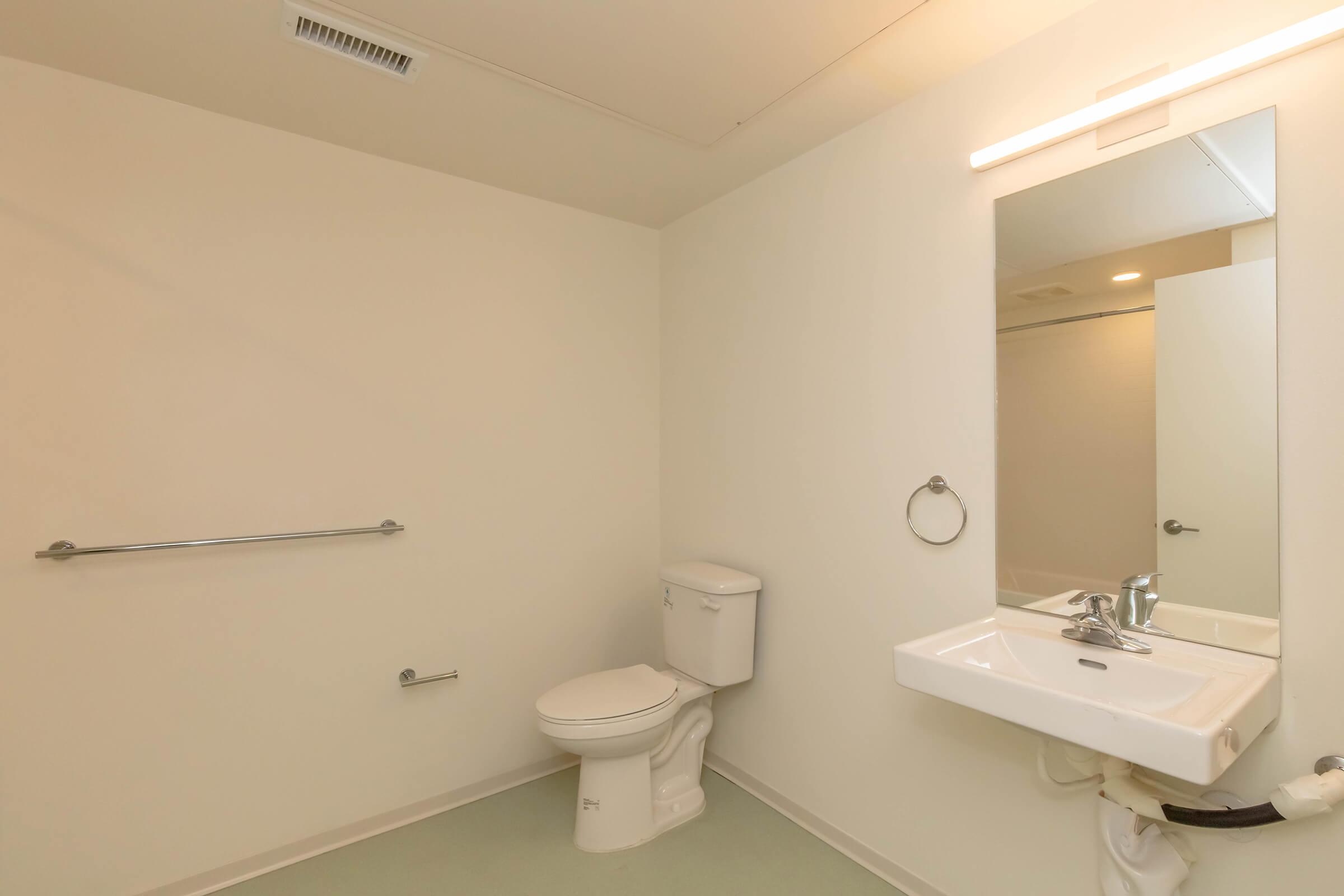 A clean, modern bathroom featuring a toilet, a small sink with a faucet, a wall-mounted mirror, and a grab bar. The walls are painted white, and the floor is a light-colored material. There is a towel ring next to the sink, and the overall design is minimalistic and functional.