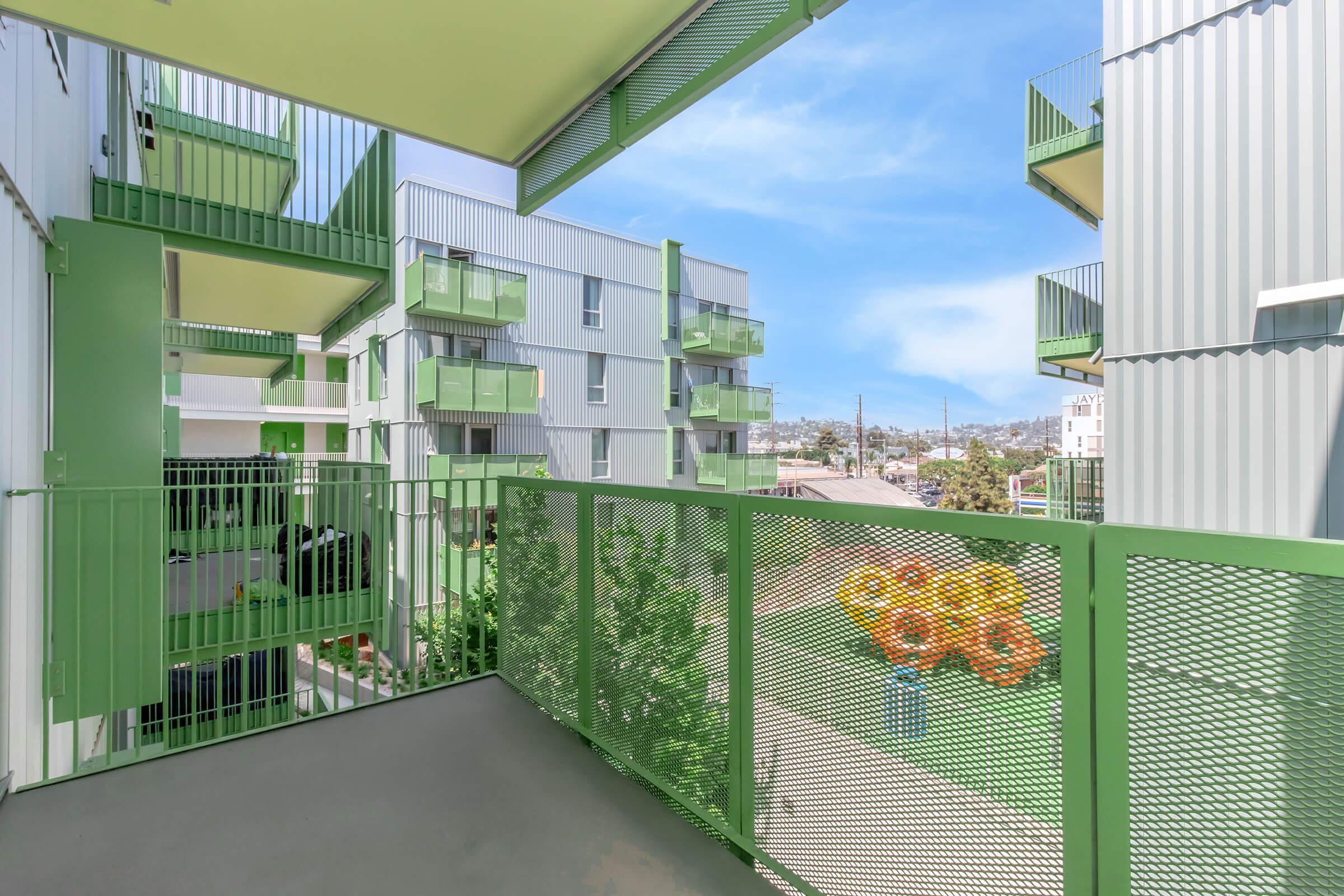 View of a modern apartment balcony featuring green railings and colorful outdoor decor. The surrounding buildings are designed with a contemporary aesthetic, showcasing a mix of green tones. Lush greenery is visible below, enhancing the urban environment with a touch of nature.