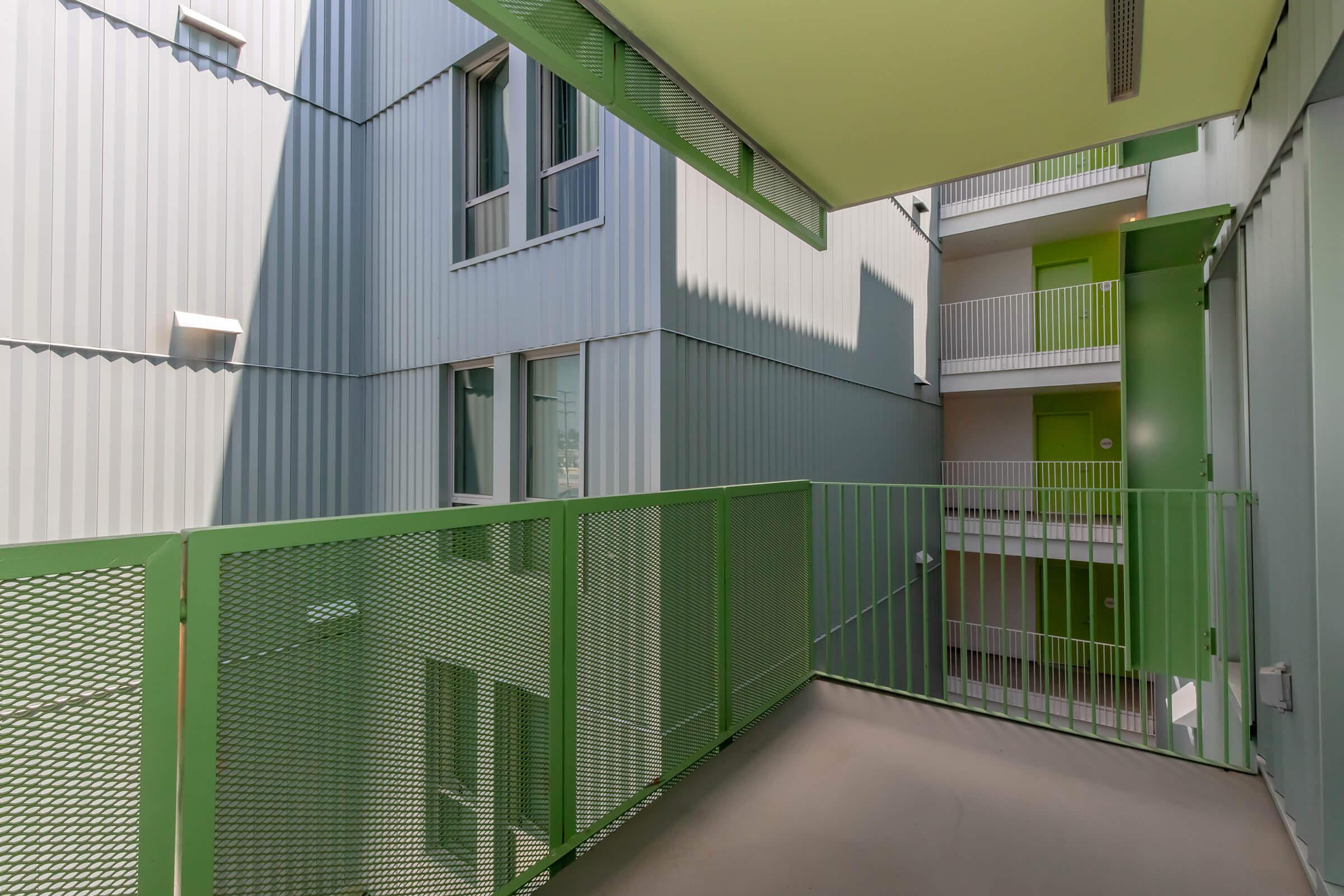 View of a modern building's interior courtyard from a balcony. The structure features grey and green metal siding with large windows. The balcony has a green metal railing and leads to an open space, showcasing a contemporary architectural design with a focus on light and airy spaces.