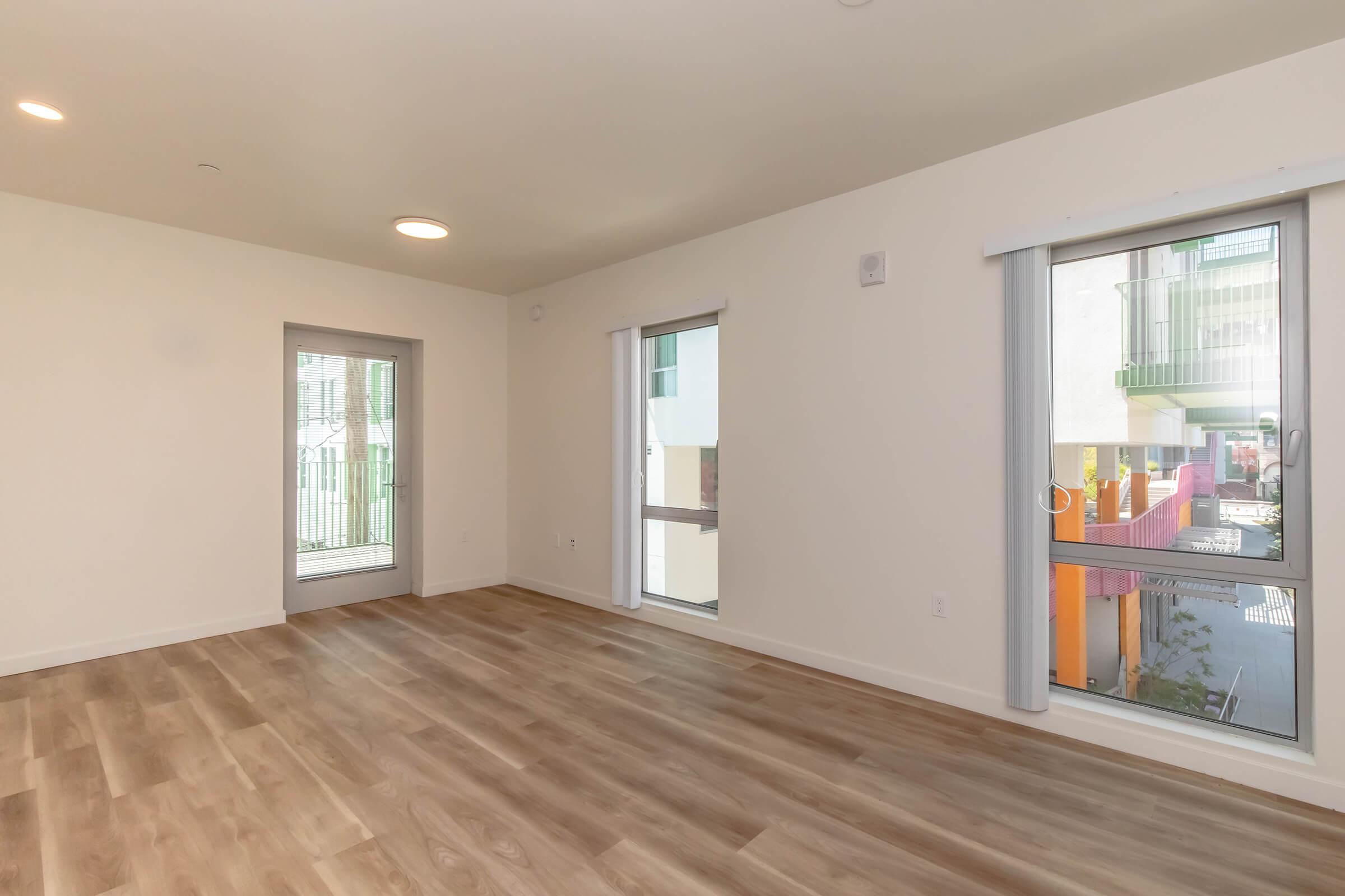 An empty room with light-colored walls and wooden flooring. Two large windows provide natural light, revealing glimpses of an exterior courtyard. The space is spacious and modern, suitable for various furnishings.