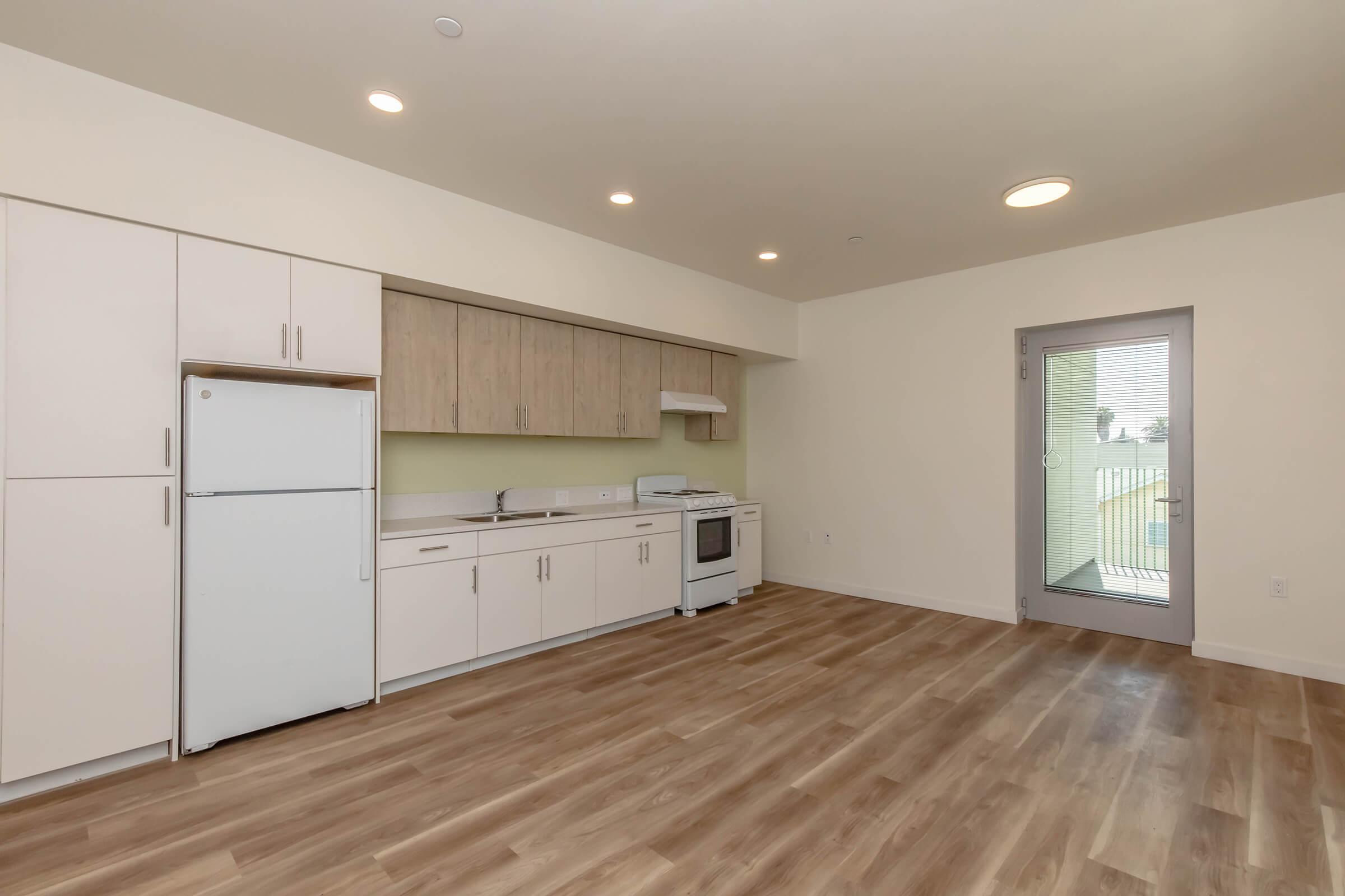 Modern kitchen in an apartment featuring white cabinetry, a refrigerator, an oven/stove, and a green accent wall. The space has laminate flooring and includes a sliding glass door that opens to a small balcony or patio area, providing natural light.