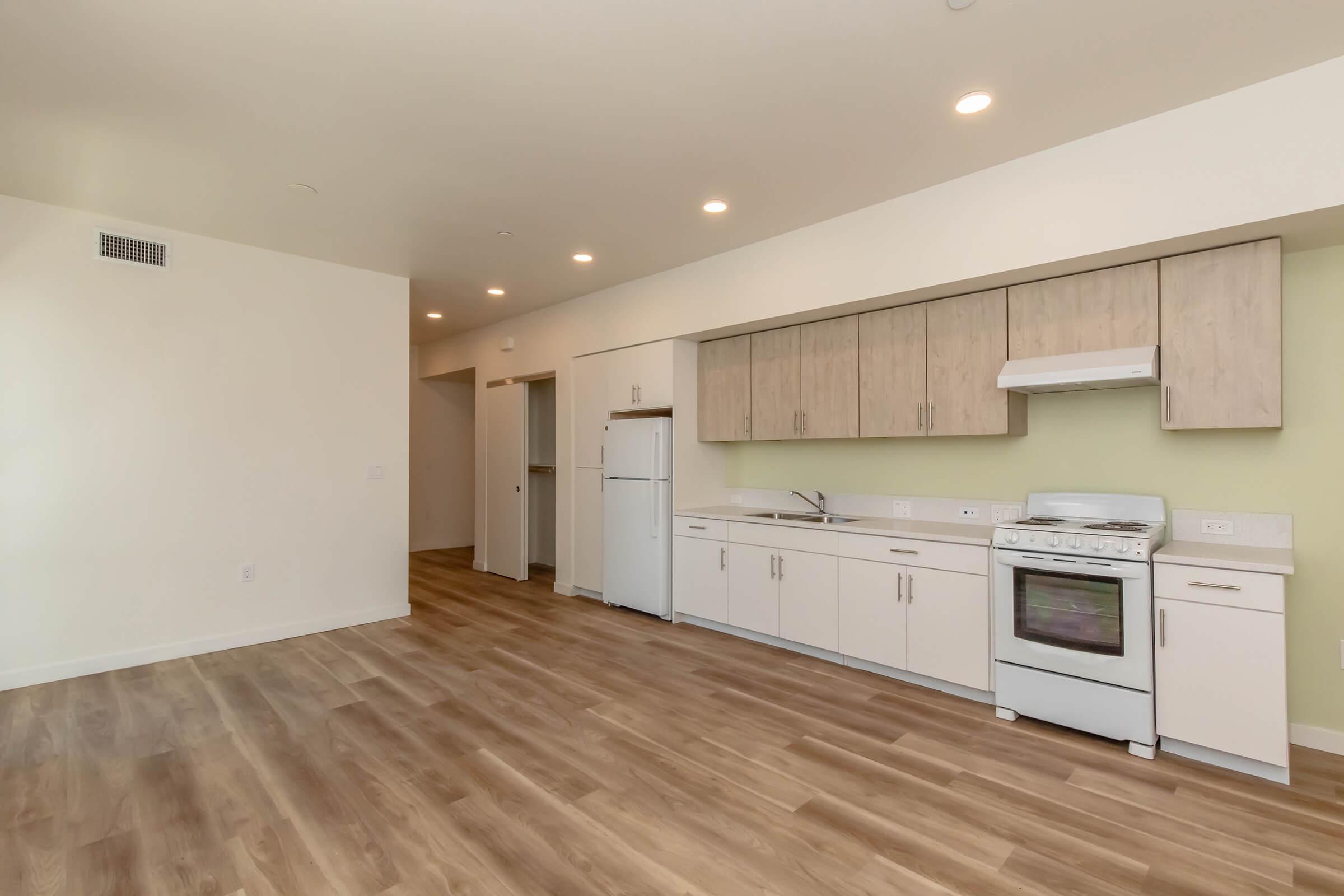 A modern, spacious kitchen and living area featuring light wood floors, white cabinetry, and a white stove. The kitchen has a refrigerator and a sink, with soft green wall accents. There are doorways leading to other rooms and ample natural light.