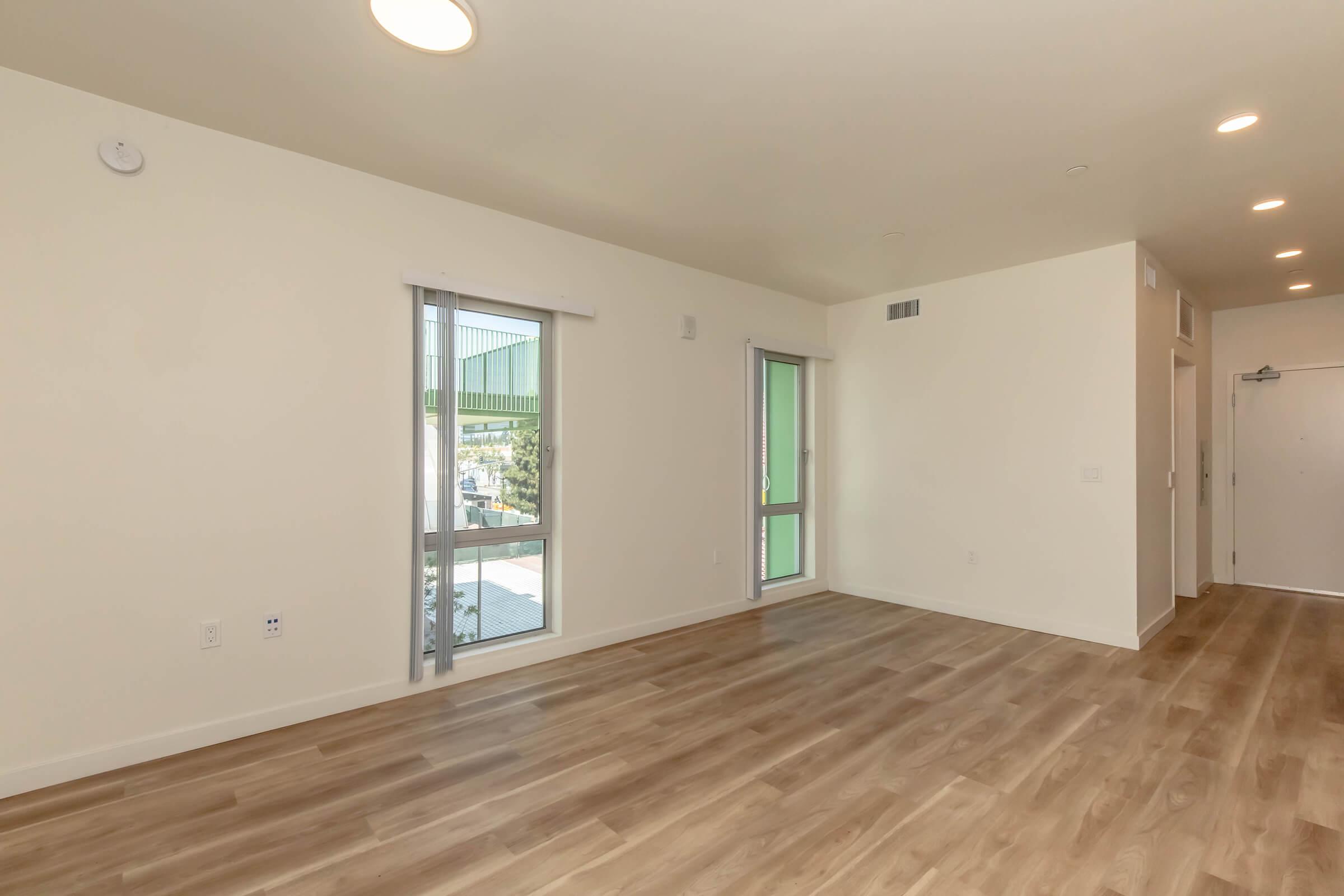 Bright and spacious interior of an empty room featuring light-colored walls, large windows with horizontal blinds, and modern laminate flooring. The layout includes an open space suitable for living or multi-purpose use, with a door leading to a balcony or patio area visible in the background.