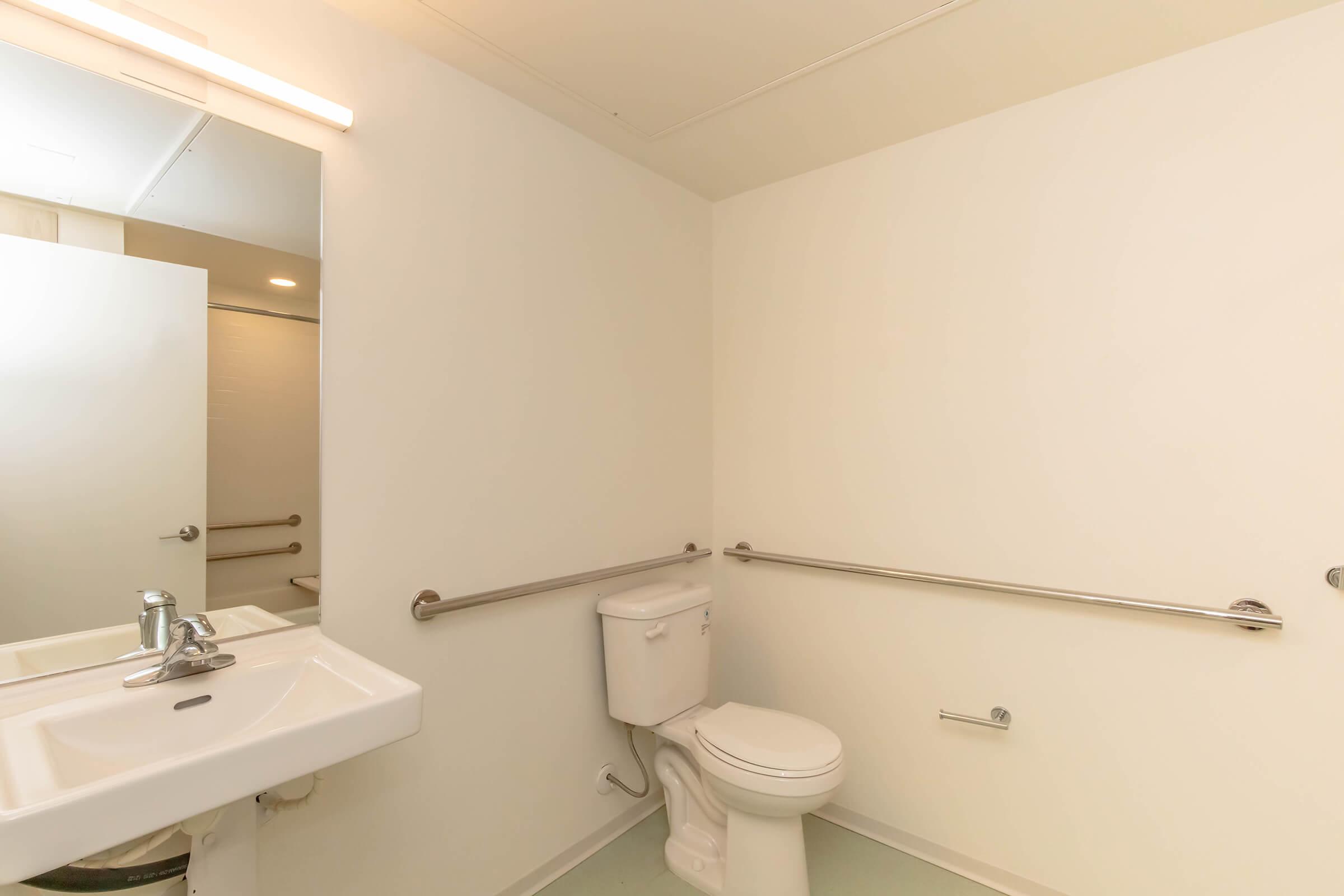 A clean, minimal restroom featuring a white wall, a toilet, a sink with a faucet, and grab bars for accessibility. A large mirror is mounted above the sink, and the space is well-lit and free of clutter.