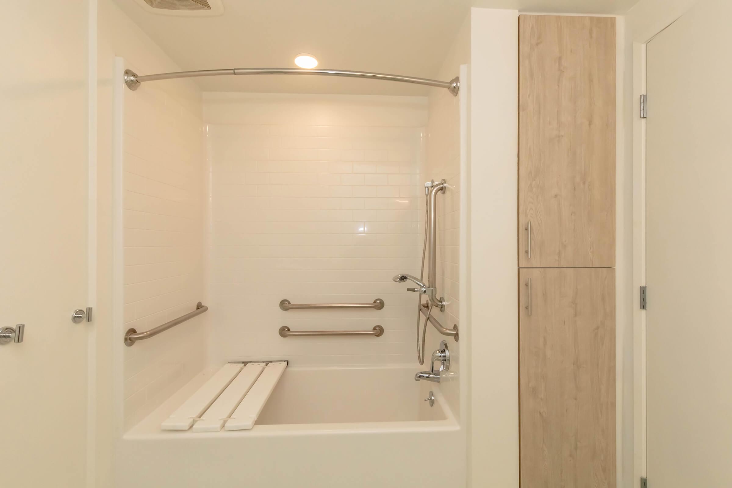 A clean and modern bathroom with a walk-in shower. The shower area features grab bars for safety, a foldable bench, and a handheld showerhead. There is a cabinetry unit for storage beside the shower, and the walls are tiled in white with a minimalist design. The overall ambiance is bright and spacious.