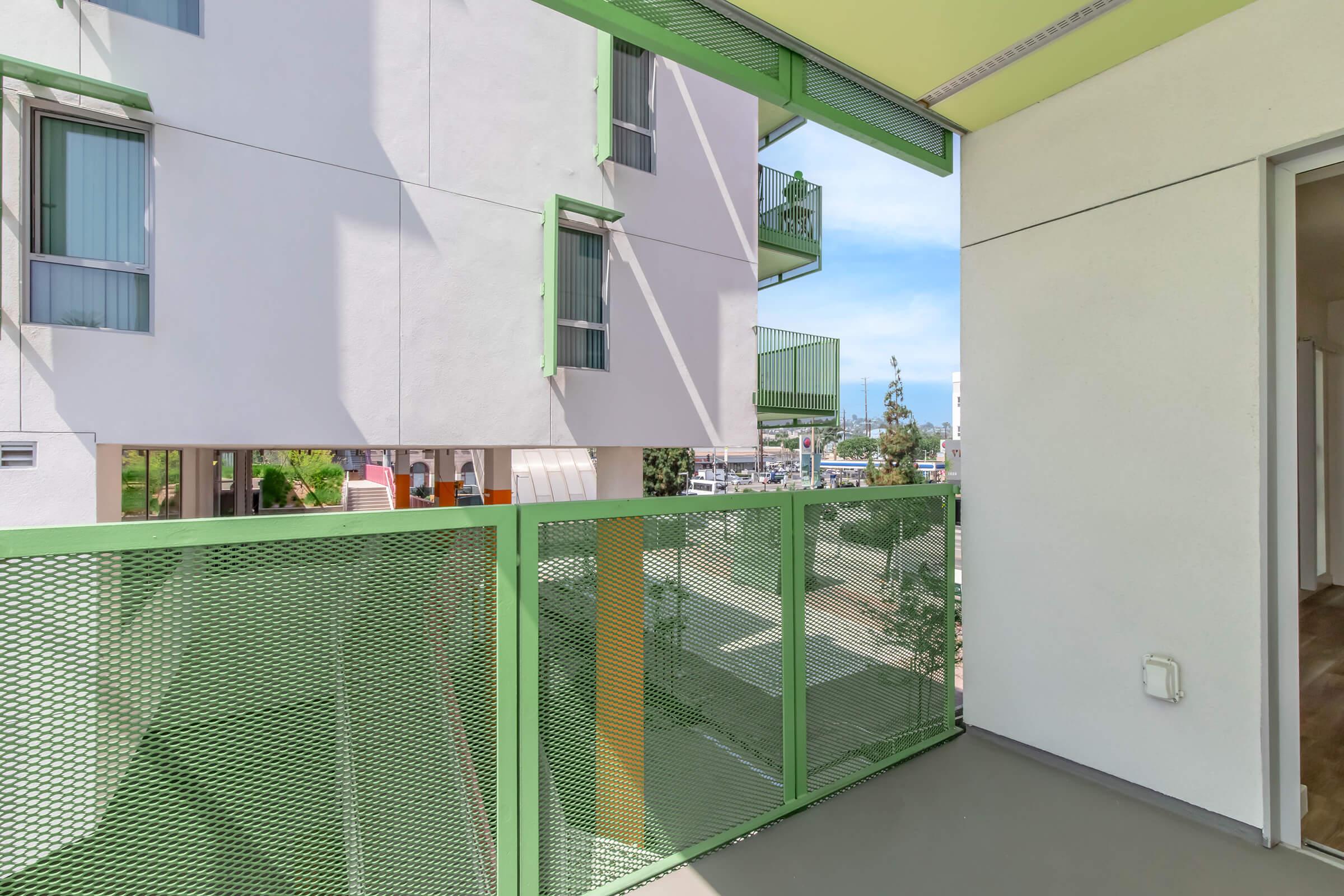 A modern balcony with green mesh railing overlooks a cityscape. The building features light-colored walls and large windows. In the background, there are nearby structures and blue sky. The scene conveys a contemporary urban living environment.