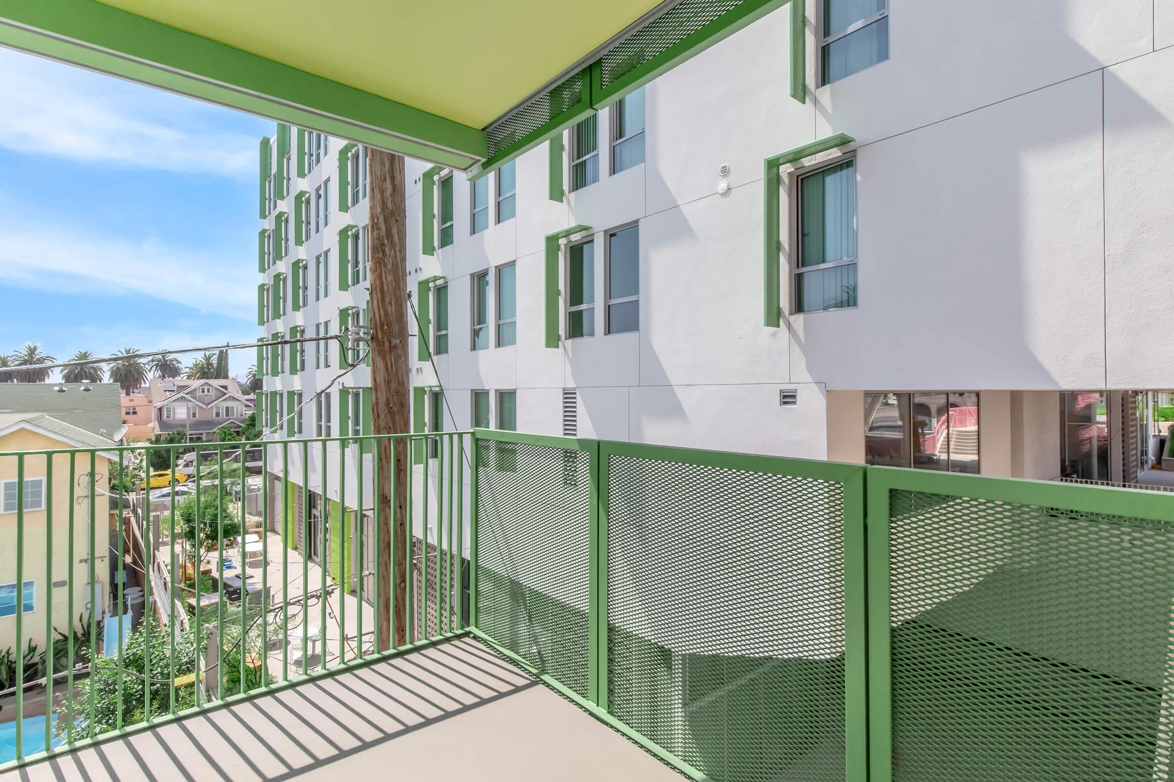 View from a balcony featuring a green railing, overlooking a courtyard with trees and neighboring buildings. The exterior of the buildings shows a modern design with large windows and green accents, set against a bright blue sky.