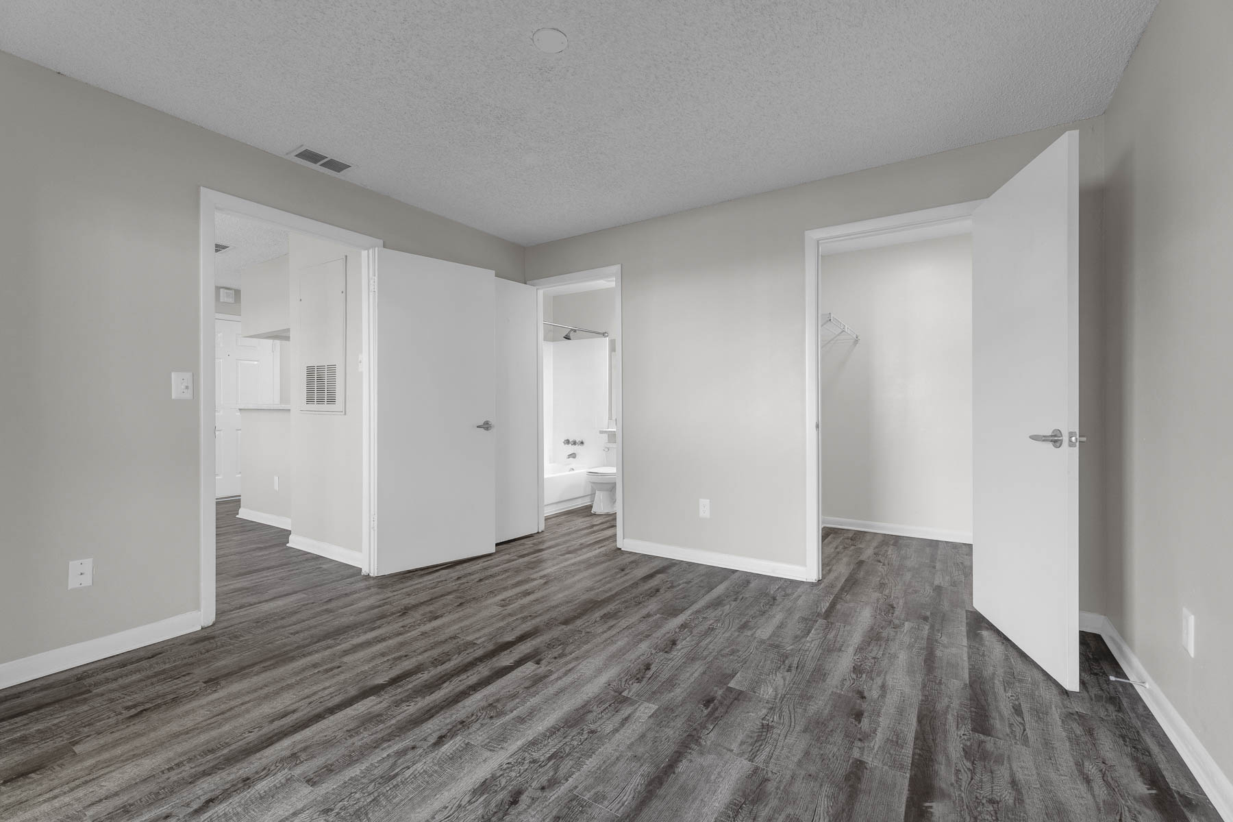 Interior view of a modern, empty room with gray walls and a wooden floor. Doorways lead to other rooms, one showing a bathroom with a tub. The space is well-lit and features white doors, creating a clean and contemporary look.