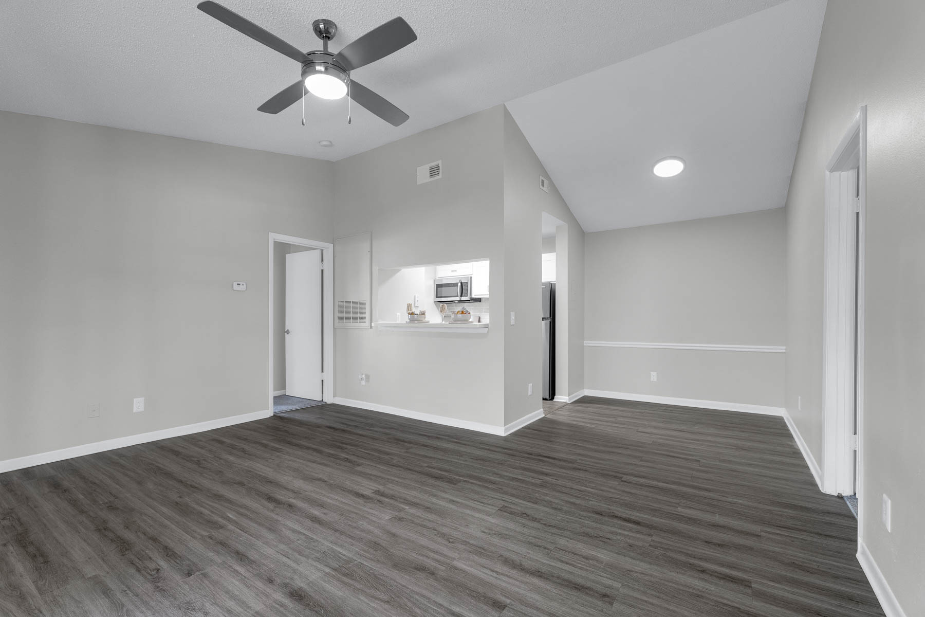 A spacious, modern living area featuring light gray walls and laminate flooring. A ceiling fan is visible, and there's an open kitchen in the background with a bar counter. The room is well-lit with natural light and has a doorway leading to another room.