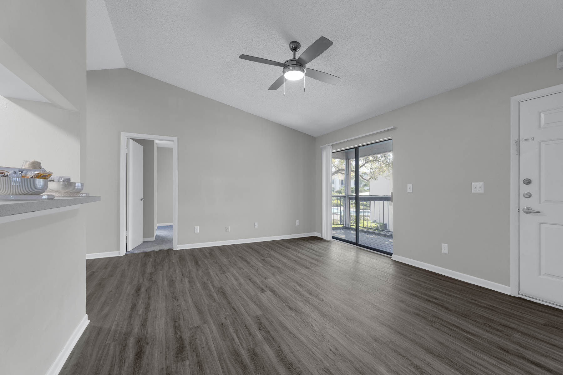 Spacious living area with a high ceiling, featuring a ceiling fan and large windows that provide natural light. The floor is covered in modern laminate flooring, and there is an open doorway leading to another room. A glass sliding door offers access to a balcony or outdoor space.