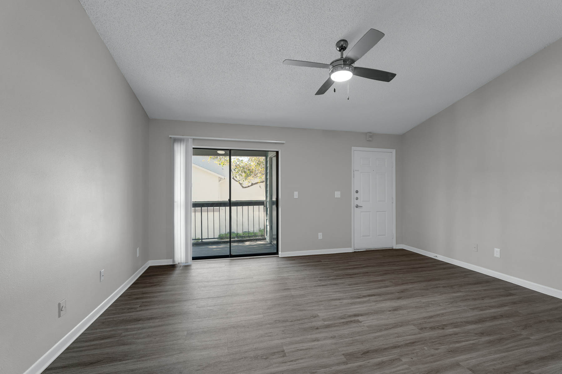 A spacious, empty living room with light gray walls and a ceiling fan. There is a large window with sliding doors leading to a balcony, letting in natural light. The floor features brown wooden laminate, and there is a front door on the right side. The overall atmosphere is airy and modern.