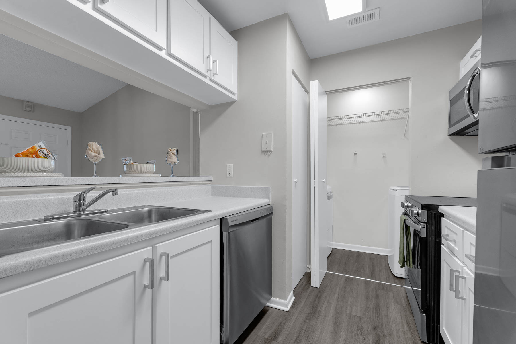 Modern kitchen featuring light-colored cabinetry, stainless steel appliances, and a double sink. The adjoining laundry area is accessible through a white door. The floor is finished with a wood-like laminate, and there is a small decorative basket of fruit on the countertop. Bright and spacious layout.
