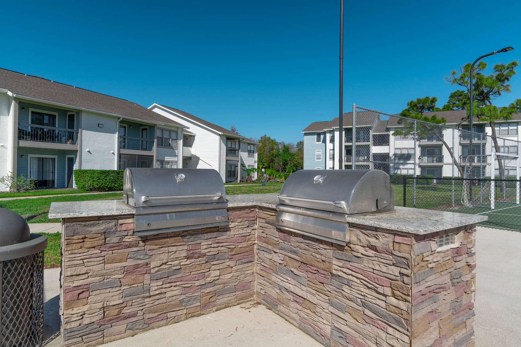 Two stainless steel grills positioned on a stone countertop, set in a park-like area near residential buildings. A tennis court is visible in the background, accompanied by clear blue skies and well-maintained greenery.