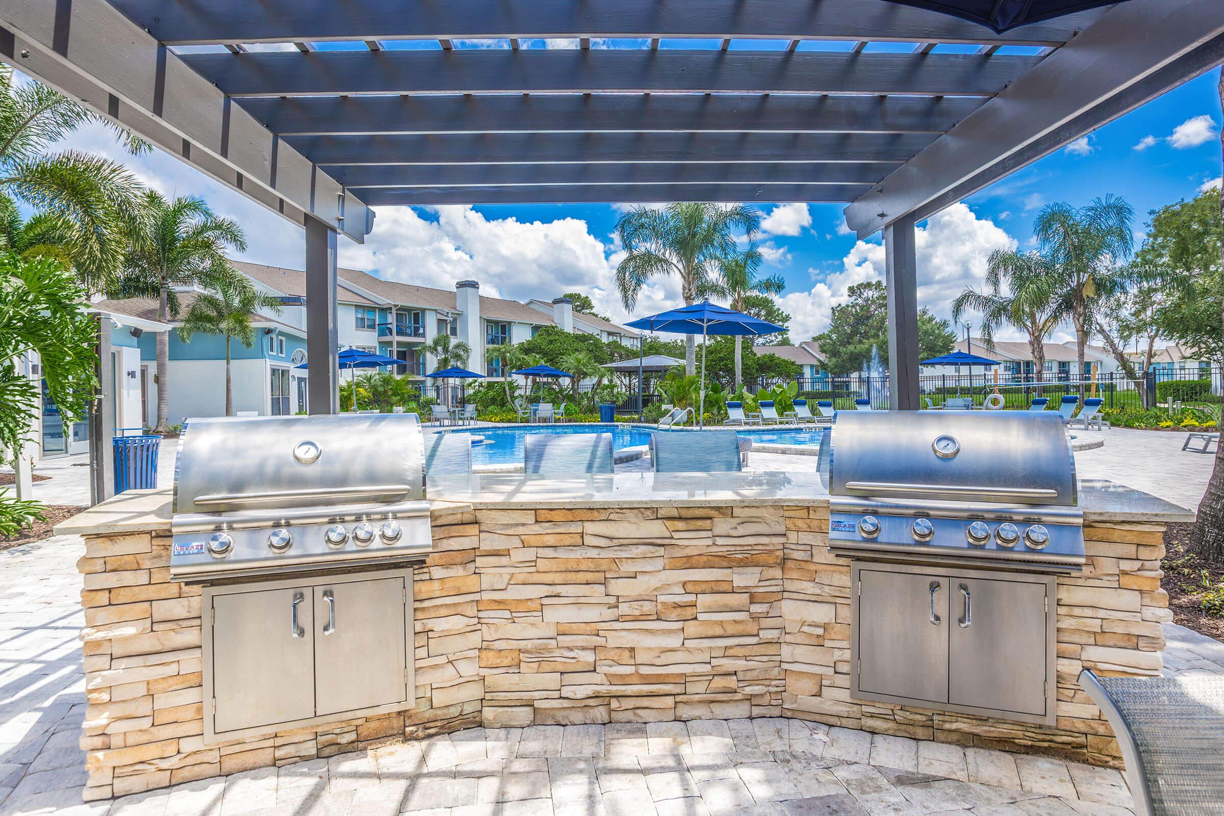 Outdoor grilling area featuring two stainless steel barbecue grills, surrounded by a stone countertop. A swimming pool is visible in the background, along with palm trees and blue sky dotted with clouds. Umbrellas and lounge chairs are arranged around the pool area, creating a relaxing atmosphere.