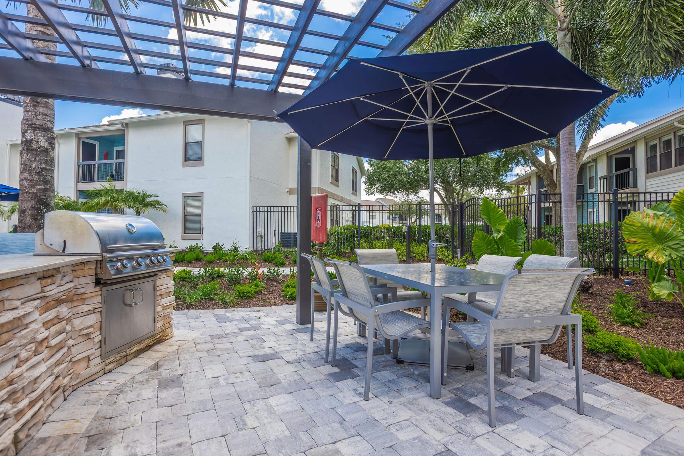 Outdoor patio area featuring a modern grill, a large umbrella over a dining table with chairs, surrounded by lush greenery and decorative plants. The setting includes a pool area and residential buildings in the background, under a clear blue sky.