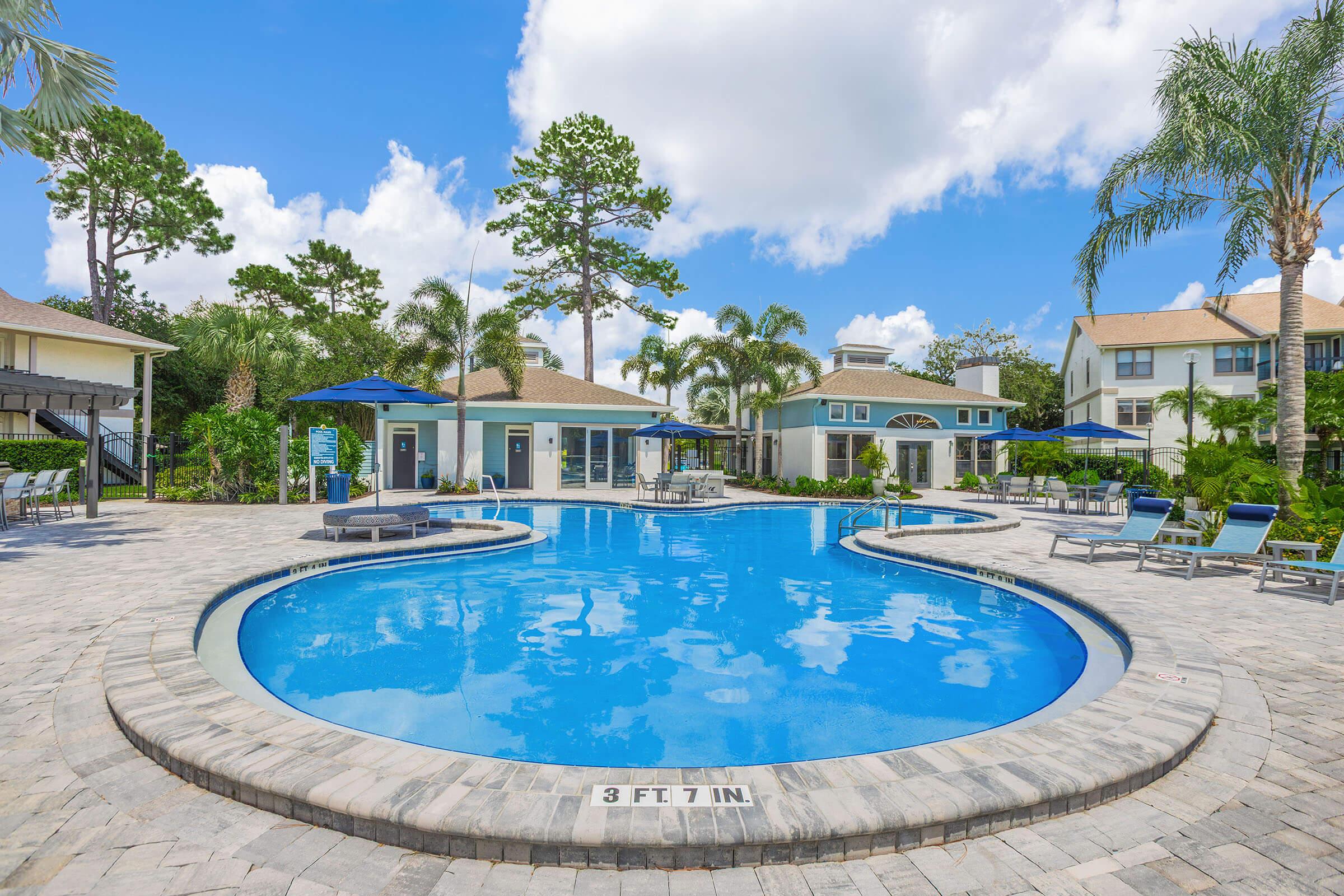 A tranquil pool area featuring a large, curved swimming pool surrounded by palm trees and lounge chairs. Nearby, there are blue umbrellas providing shade, and a clubhouse with large windows is visible in the background under a bright blue sky with fluffy white clouds.