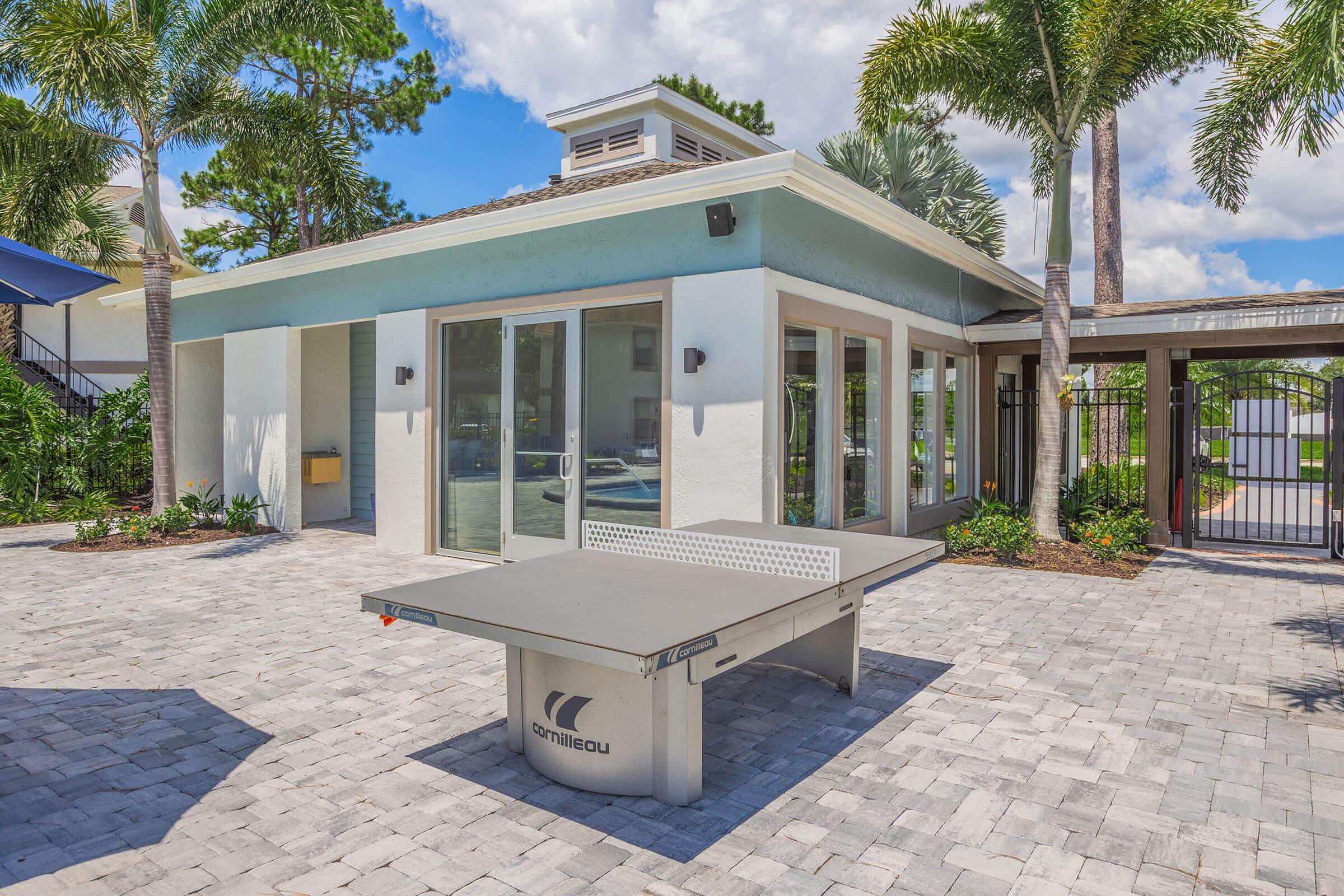 A ping pong table is set up in an outdoor area surrounded by palm trees and a well-maintained landscape, with a modern building featuring large glass doors in the background. The scene is bright and sunny, creating a welcoming atmosphere for recreational activities.