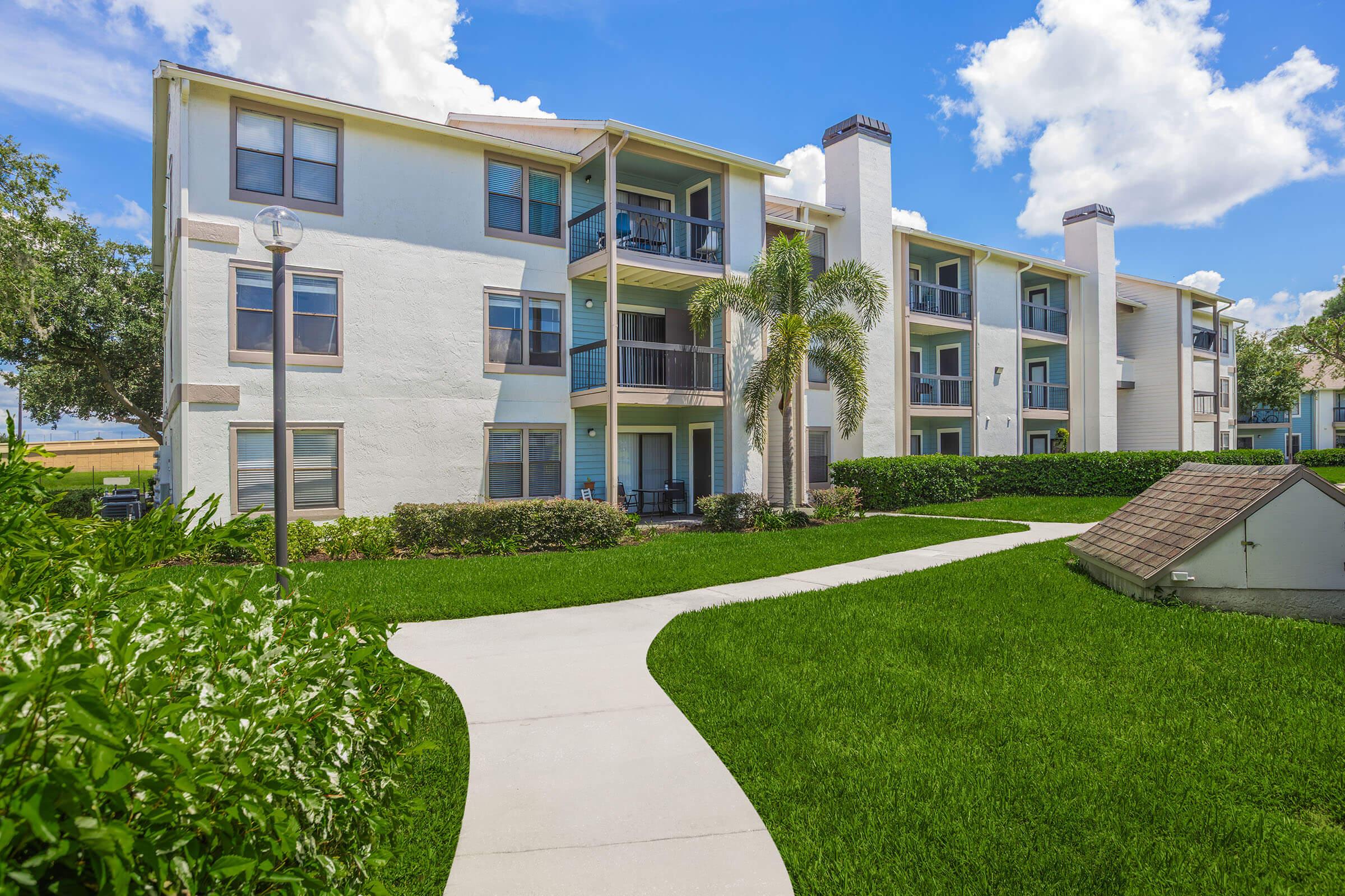 A well-maintained apartment building featuring a white exterior with multiple balconies. Green landscaping surrounds the building, with a paved path leading to the entrance. The sky is blue with fluffy clouds, creating a bright and inviting atmosphere.