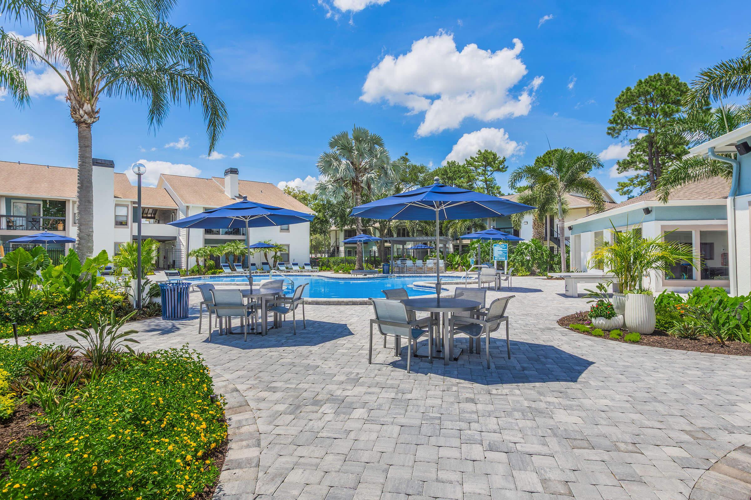 A sunny outdoor area featuring a swimming pool surrounded by lush greenery, patio tables, and blue umbrellas. The landscape includes a stone-paved walkway and vibrant flowers, complemented by palm trees under a bright blue sky with scattered clouds. Ideal for relaxation and leisure.