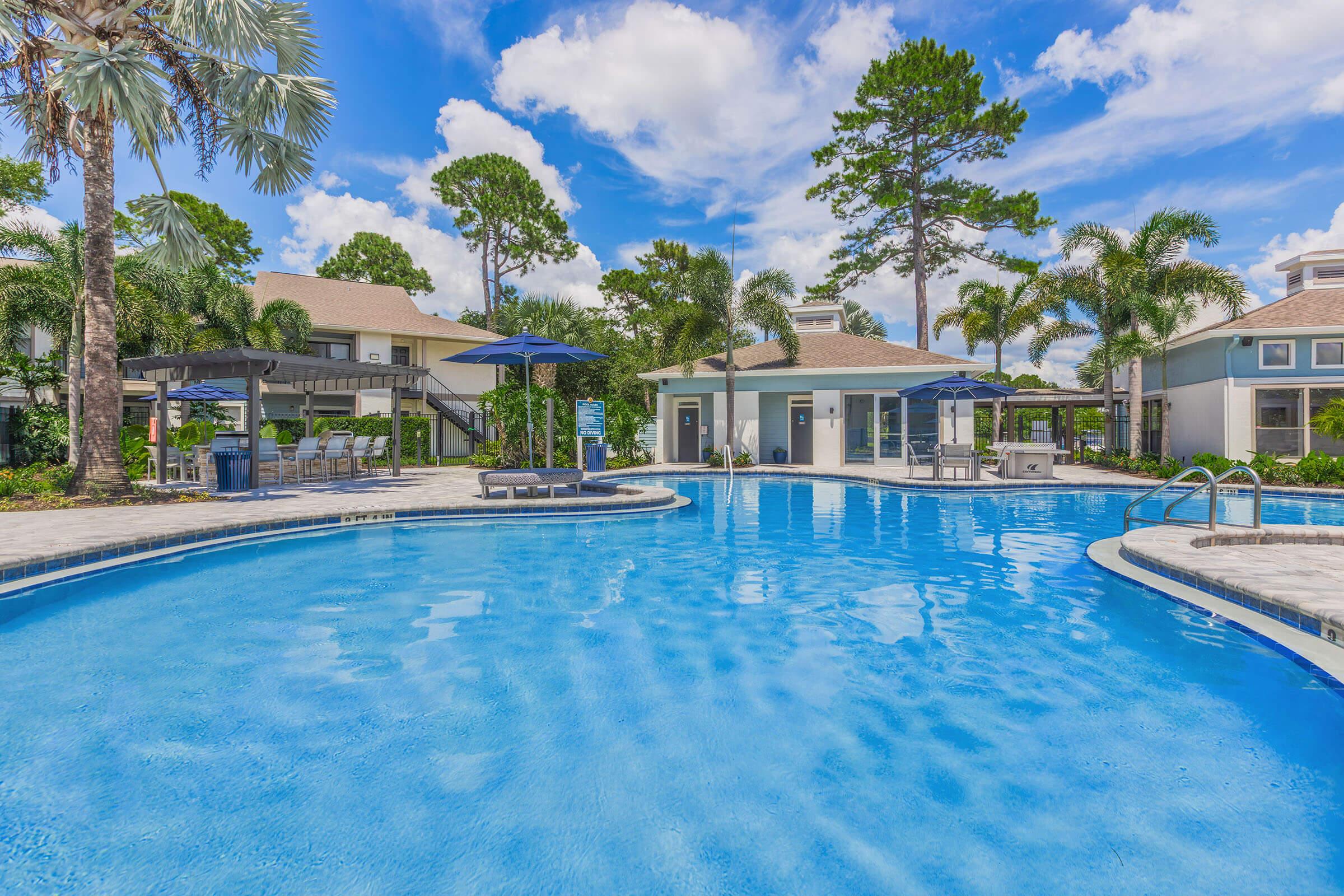 A tranquil outdoor pool area surrounded by palm trees and greenery, featuring lounge chairs and umbrellas. A clubhouse with large windows is visible in the background, set against a bright blue sky dotted with fluffy white clouds. The scene conveys a relaxing, resort-like atmosphere.