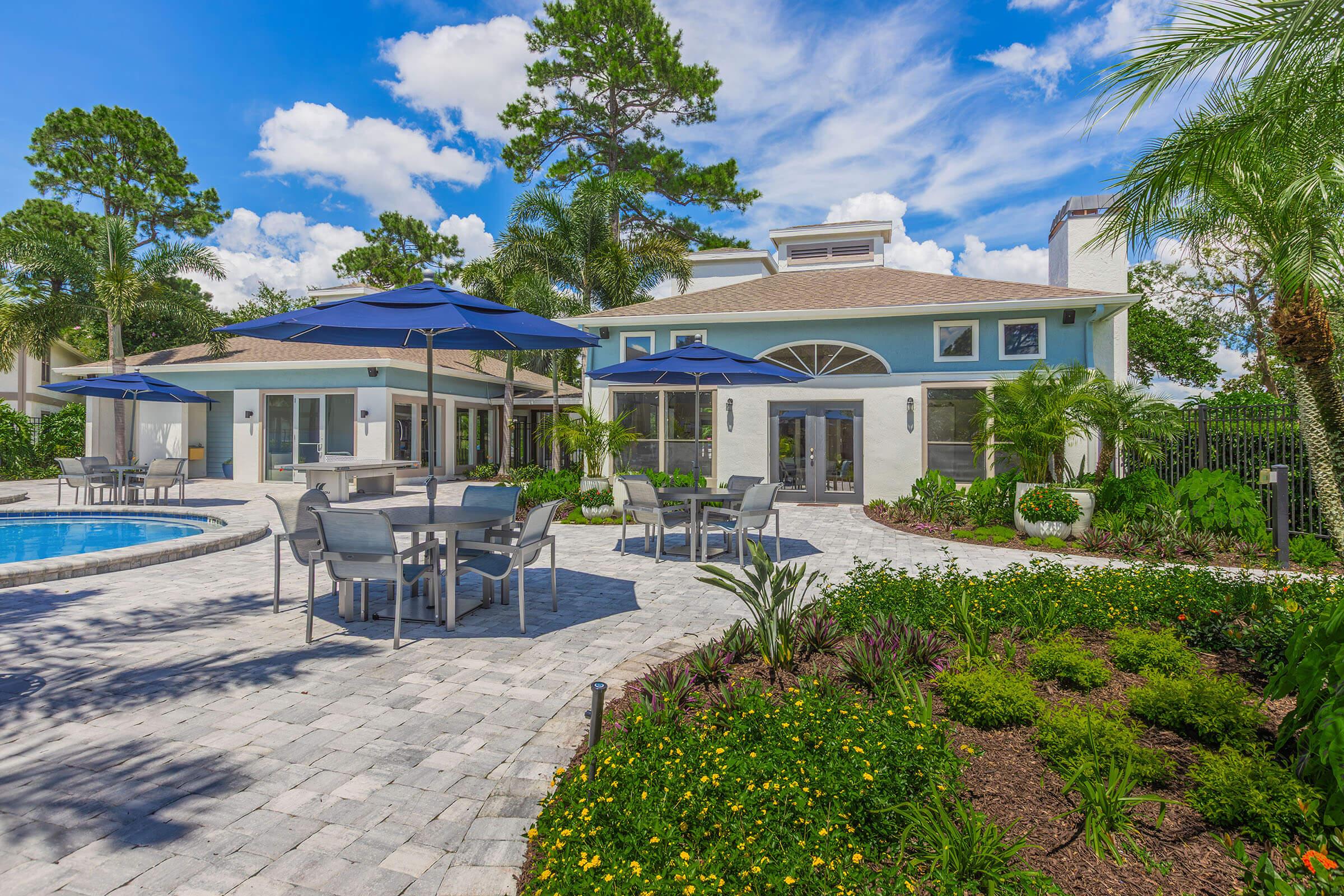 A scenic view of a modern backyard featuring a swimming pool, gray stone patio, and several outdoor dining tables with blue umbrellas. Lush greenery and vibrant flowers surround the area, complemented by tall palm trees and a blue sky with fluffy clouds in the background.