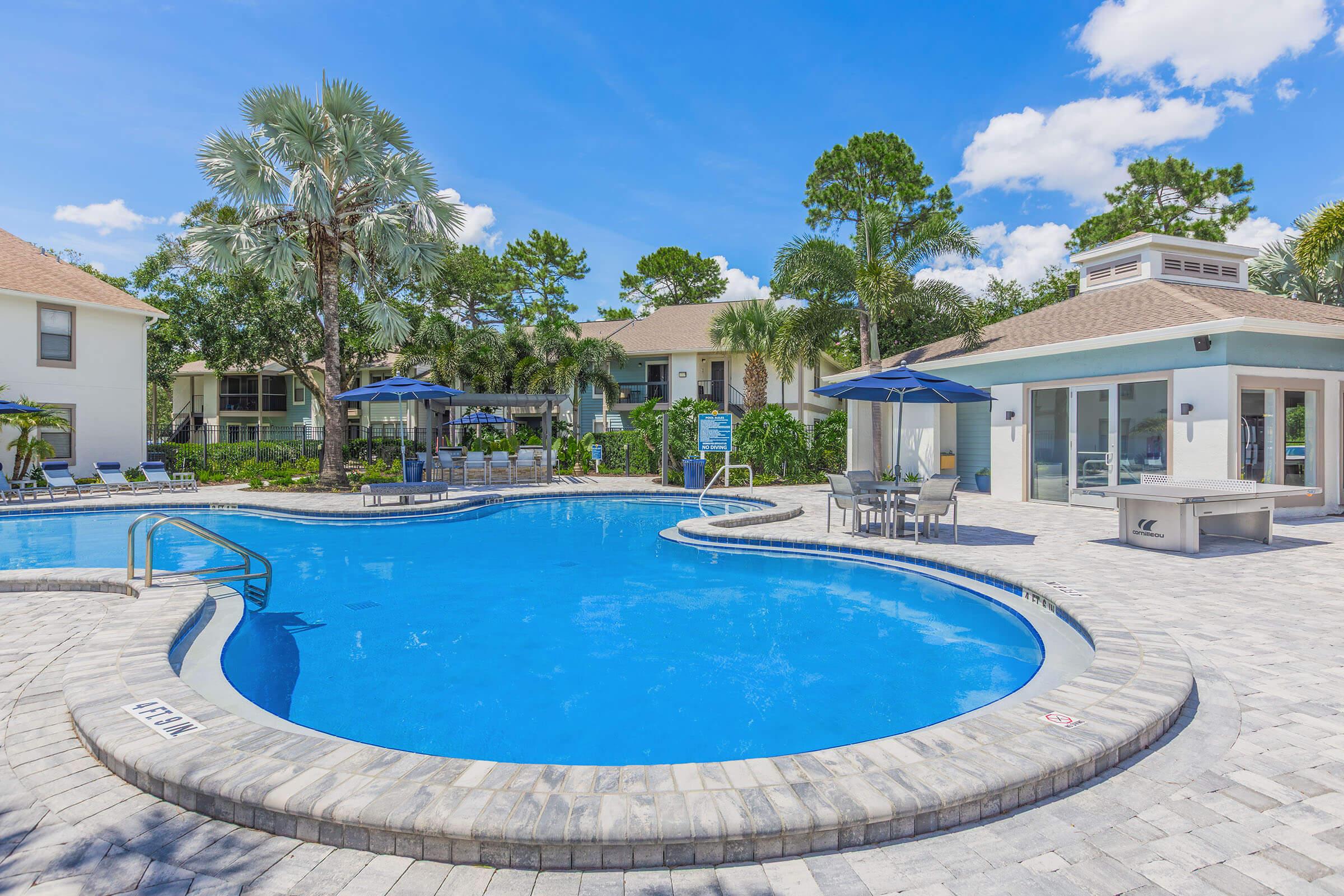 A sunny outdoor pool area featuring a curved swimming pool with clear blue water, surrounded by lounge chairs and shaded umbrellas. Lush greenery and palm trees add a tropical vibe, with a nearby community building visible. The bright sky and well-maintained landscape create a relaxing atmosphere.