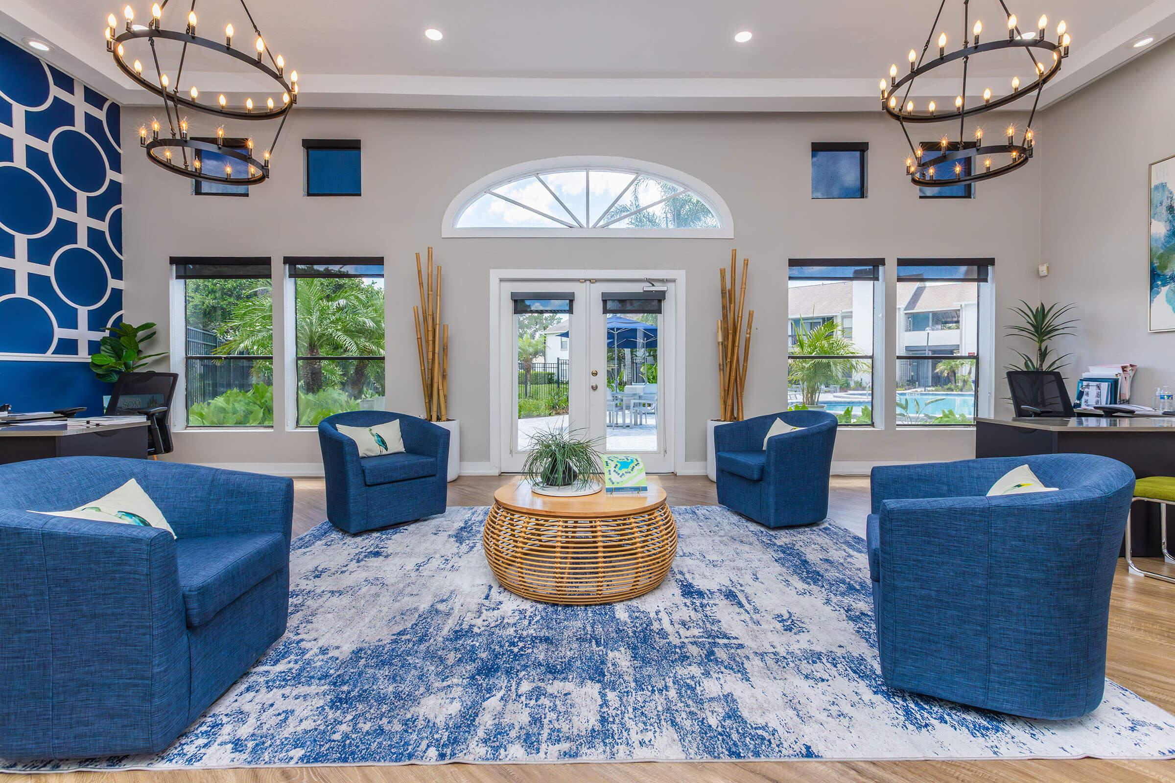 A modern lounge area featuring four blue upholstered chairs arranged around a round wicker coffee table on a blue and white patterned rug. The space has large windows allowing natural light, decorative plants, and an elegant chandelier overhead, with a stylish accent wall displaying circular patterns.