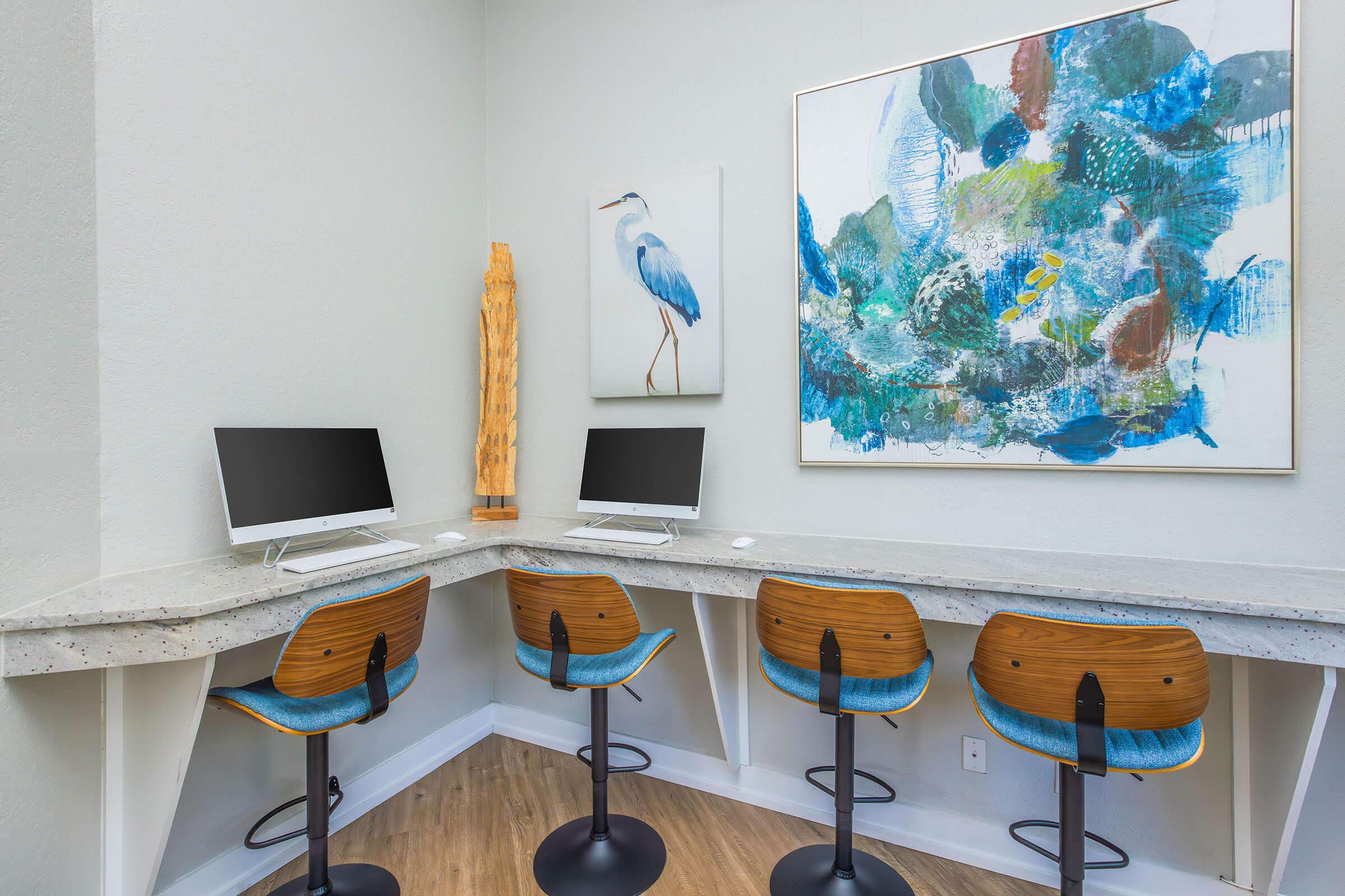 A modern workspace featuring three wooden barstools at a sleek counter with two computers. The wall is adorned with an abstract painting and a framed artwork of a blue heron. The room has light-colored walls and wooden flooring, creating a contemporary and inviting atmosphere.