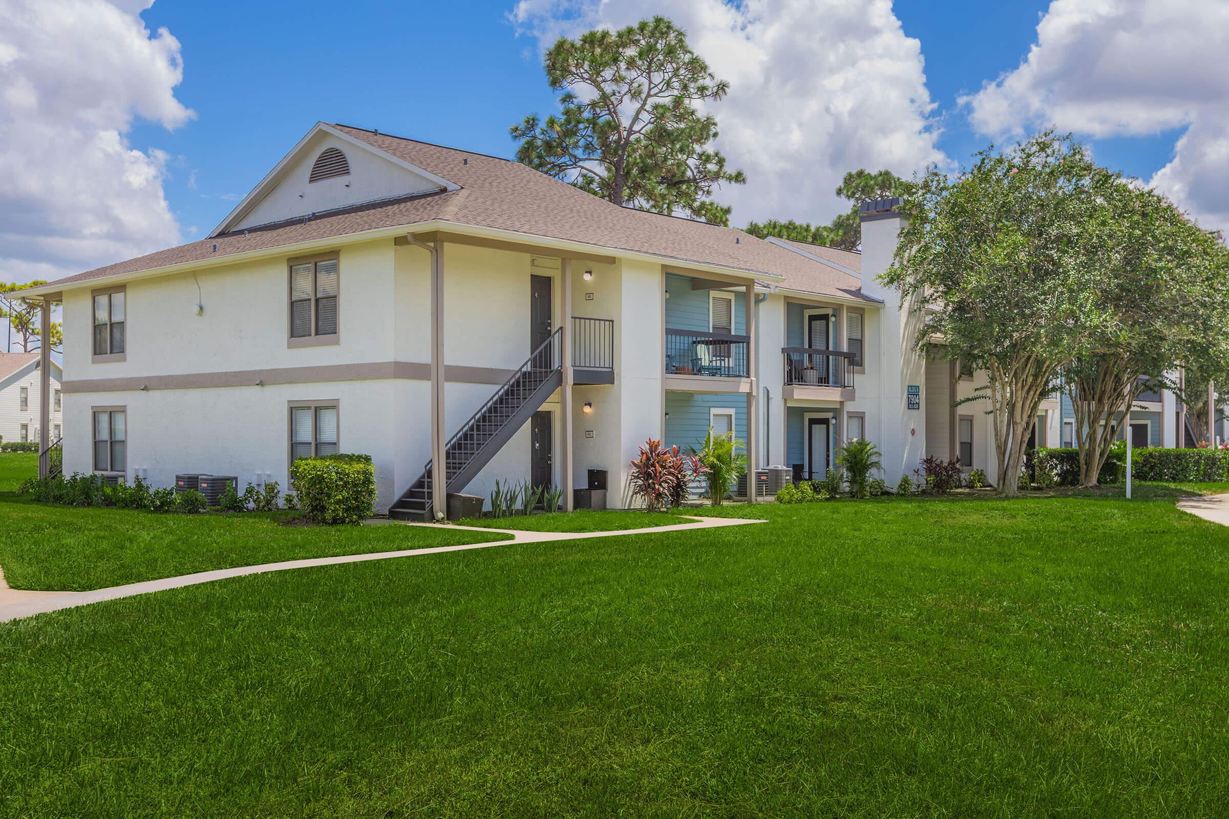 A scenic view of a residential apartment complex featuring two-story buildings. The buildings are surrounded by well-maintained green lawns and landscaped shrubs. There are walkways leading to the entrances, with a backdrop of blue skies and fluffy clouds.