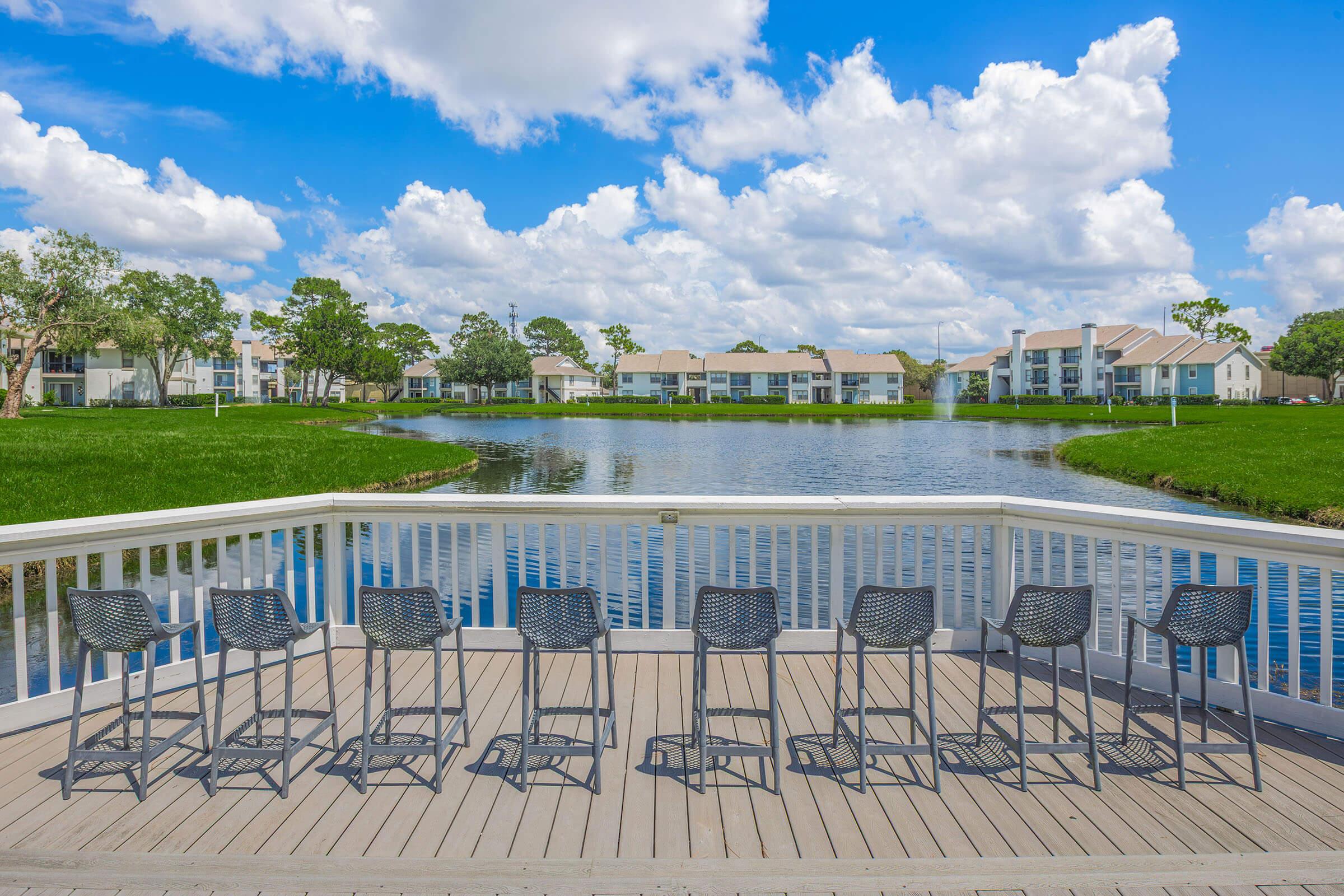 View from a deck overlooking a serene lake surrounded by lush greenery and residential buildings. The foreground features a row of gray outdoor bar stools lined up along the deck, while fluffy white clouds drift across the bright blue sky.
