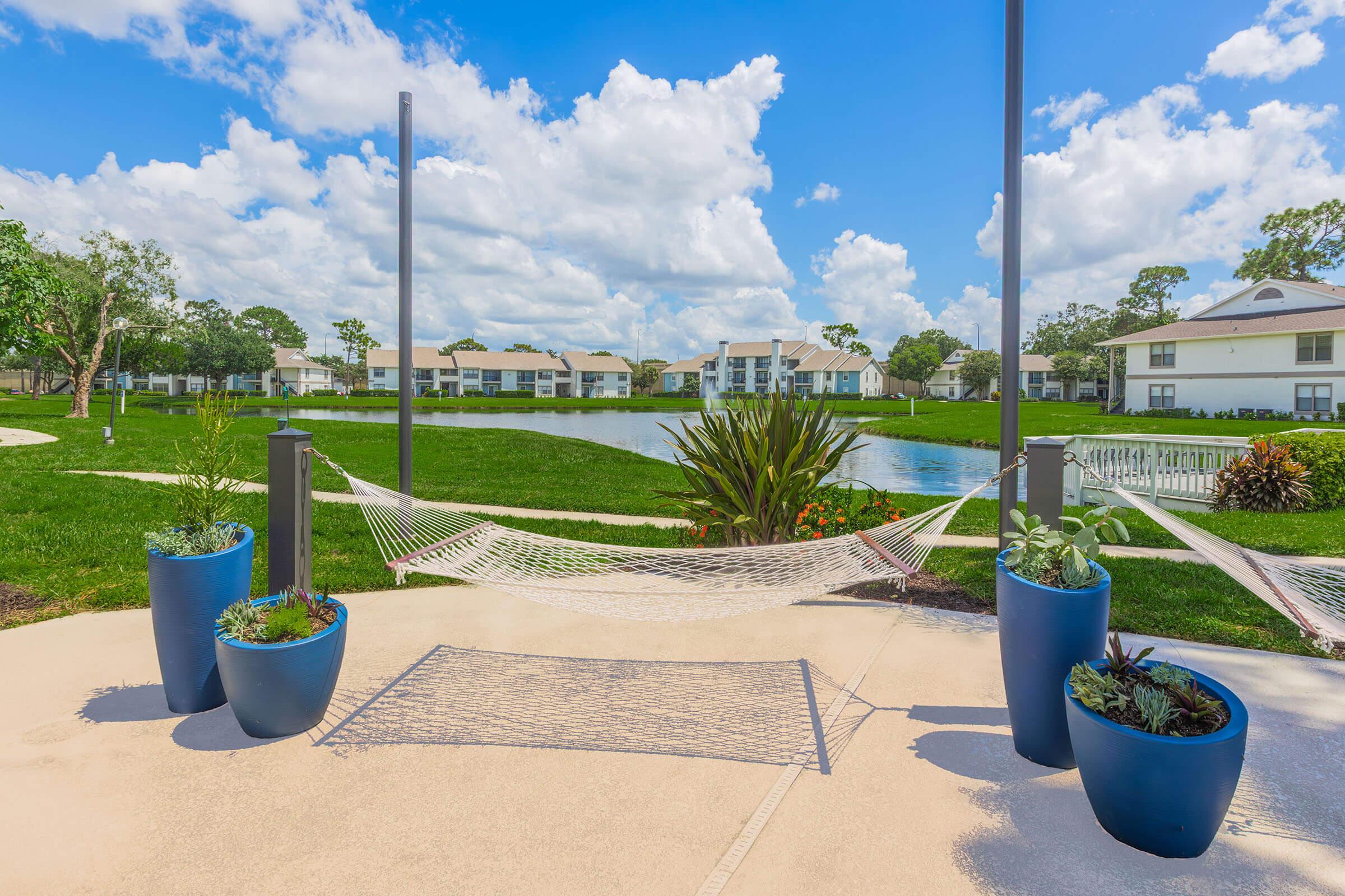 A serene outdoor space featuring a hammock between two posts, surrounded by potted plants. In the background, a peaceful pond and residential buildings under a bright blue sky with fluffy white clouds create a relaxing atmosphere.