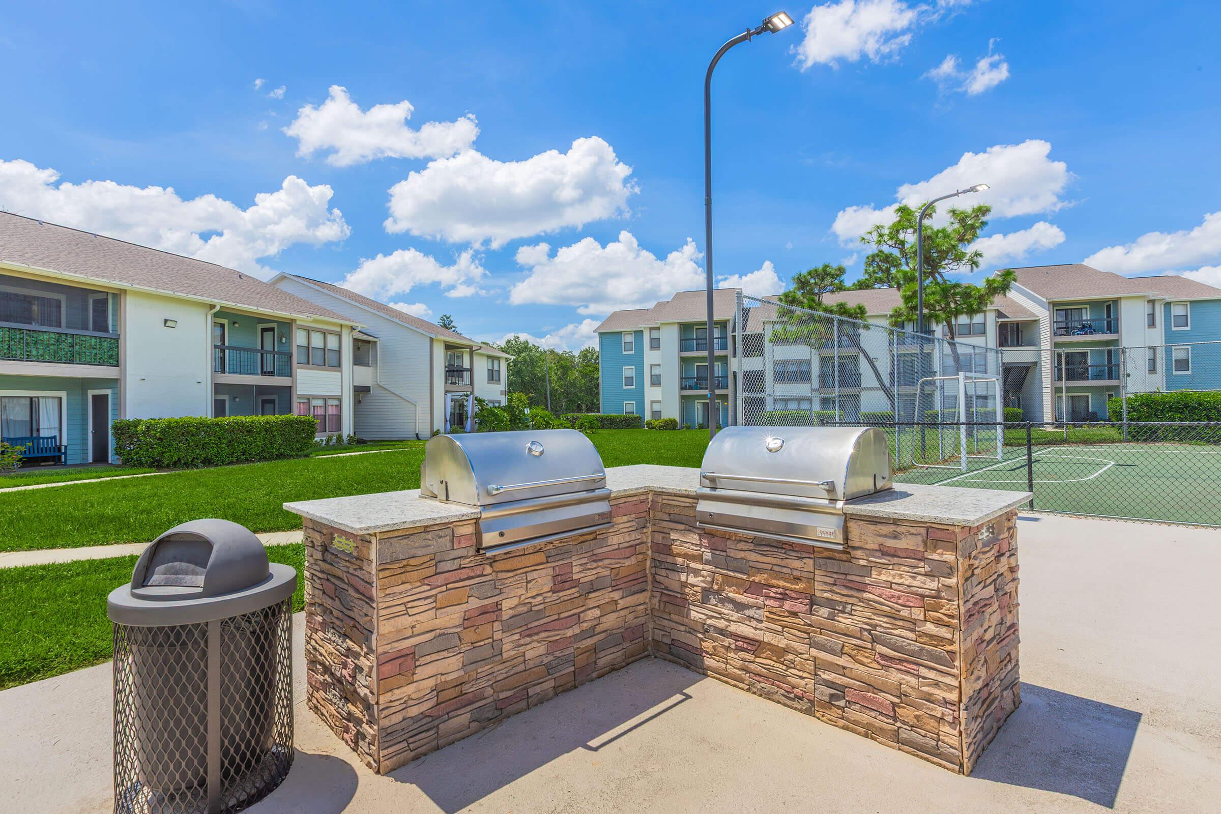 Outdoor grilling area featuring two stainless steel barbecue grills with a stone surround, situated in a landscaped area. In the background, there are apartment buildings and a sports court under a bright blue sky with fluffy white clouds. A trash can is also visible nearby.