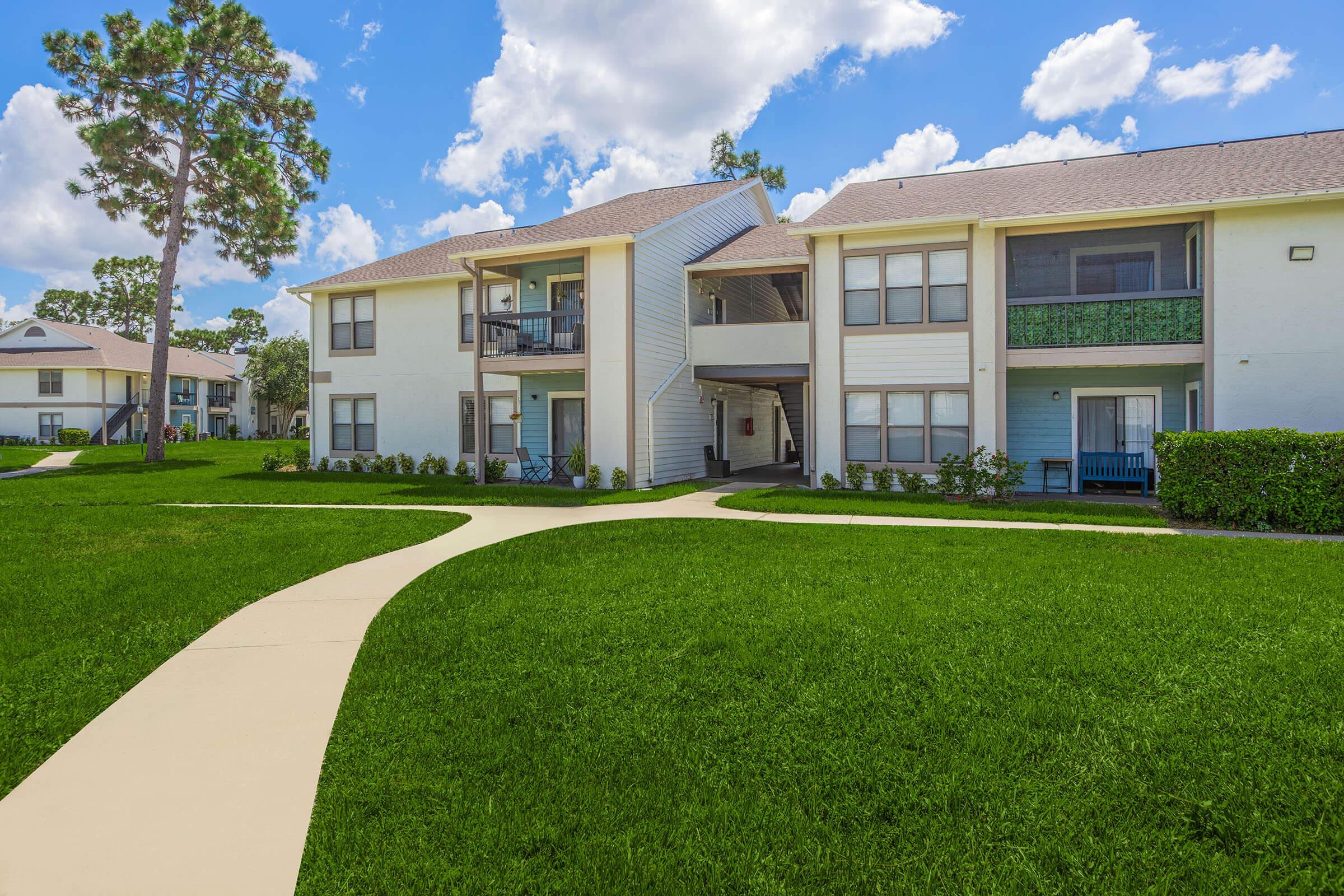 A well-maintained apartment complex featuring two buildings with balconies, surrounded by lush green lawns and neatly paved pathways. The sky is bright blue with fluffy clouds, creating a pleasant outdoor atmosphere.