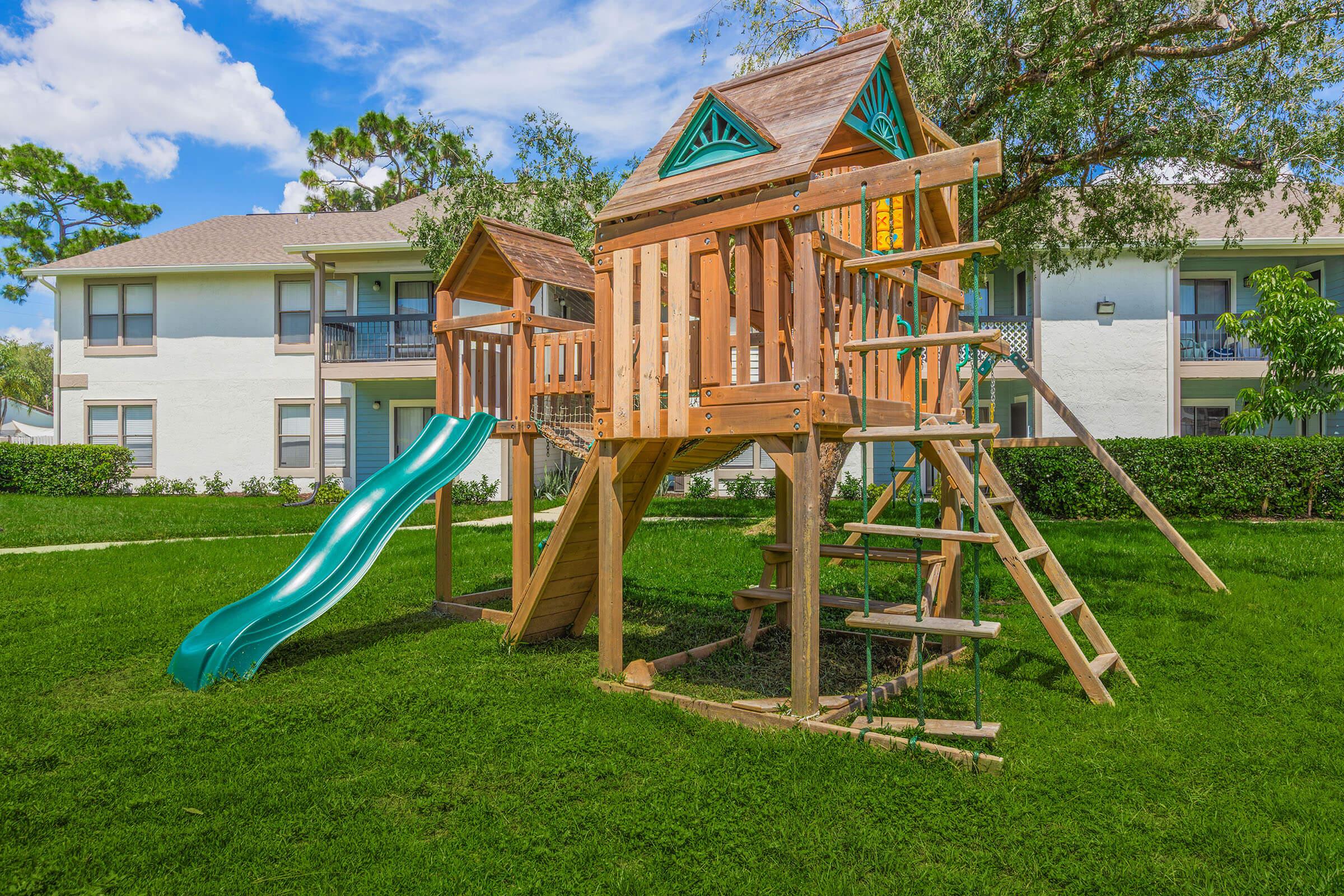 Wooden playground structure in a green backyard featuring a slide, a climbing ladder, and two levels. The playhouse has a decorative roof and is surrounded by grass and small trees, with an apartment building visible in the background under a clear blue sky.