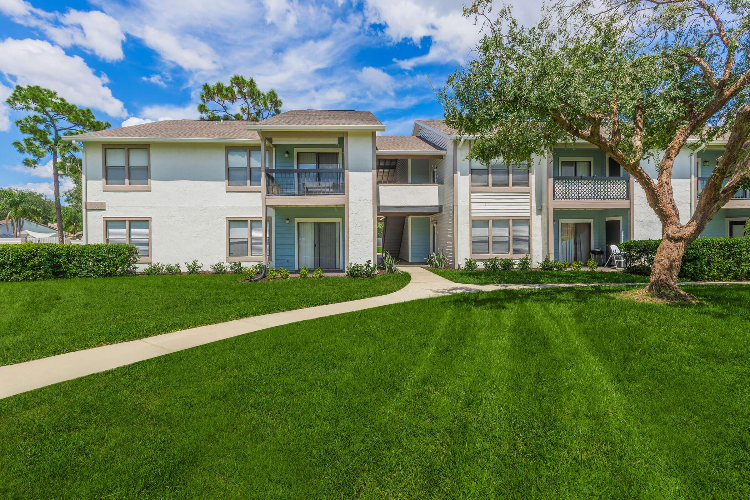 A well-maintained residential apartment complex with two-story buildings. The buildings feature balconies and are surrounded by lush green grass, shaded by trees under a bright blue sky with scattered white clouds. A paved walkway leads through the manicured lawn.