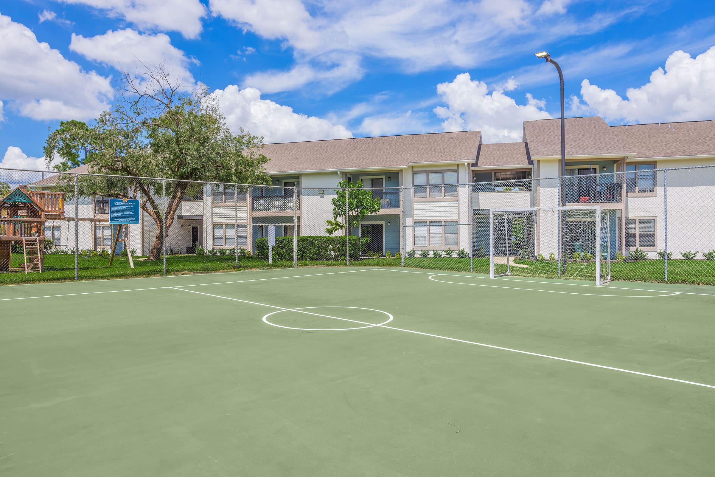 A clean, empty basketball court with a soccer goal, surrounded by a green lawn and a residential building in the background. The sky is bright with fluffy white clouds, and there is a playground visible behind the court, enclosed by a fence.