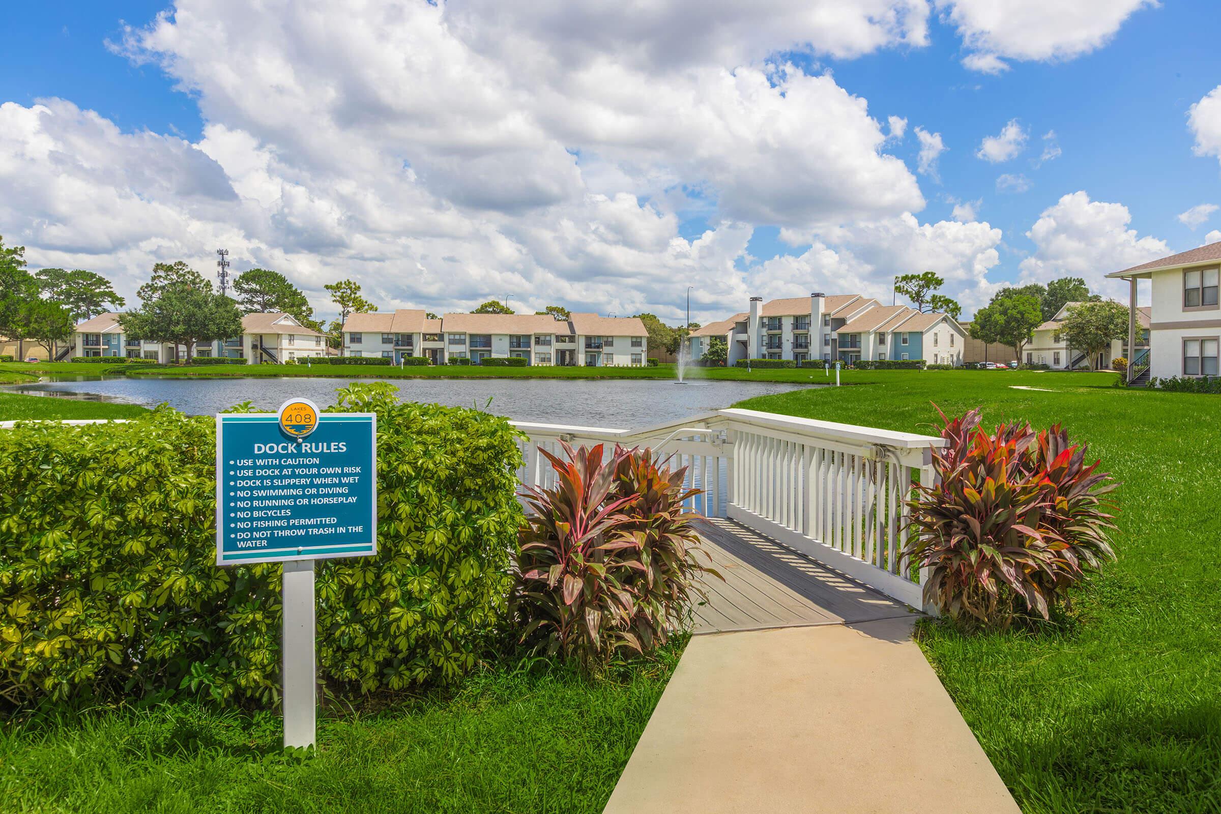 A scenic view of a tranquil lake with a sign displaying "Dock Rules" on the left. The path leads to a wooden dock, surrounded by colorful landscaping and residential buildings in the background under a partly cloudy sky.