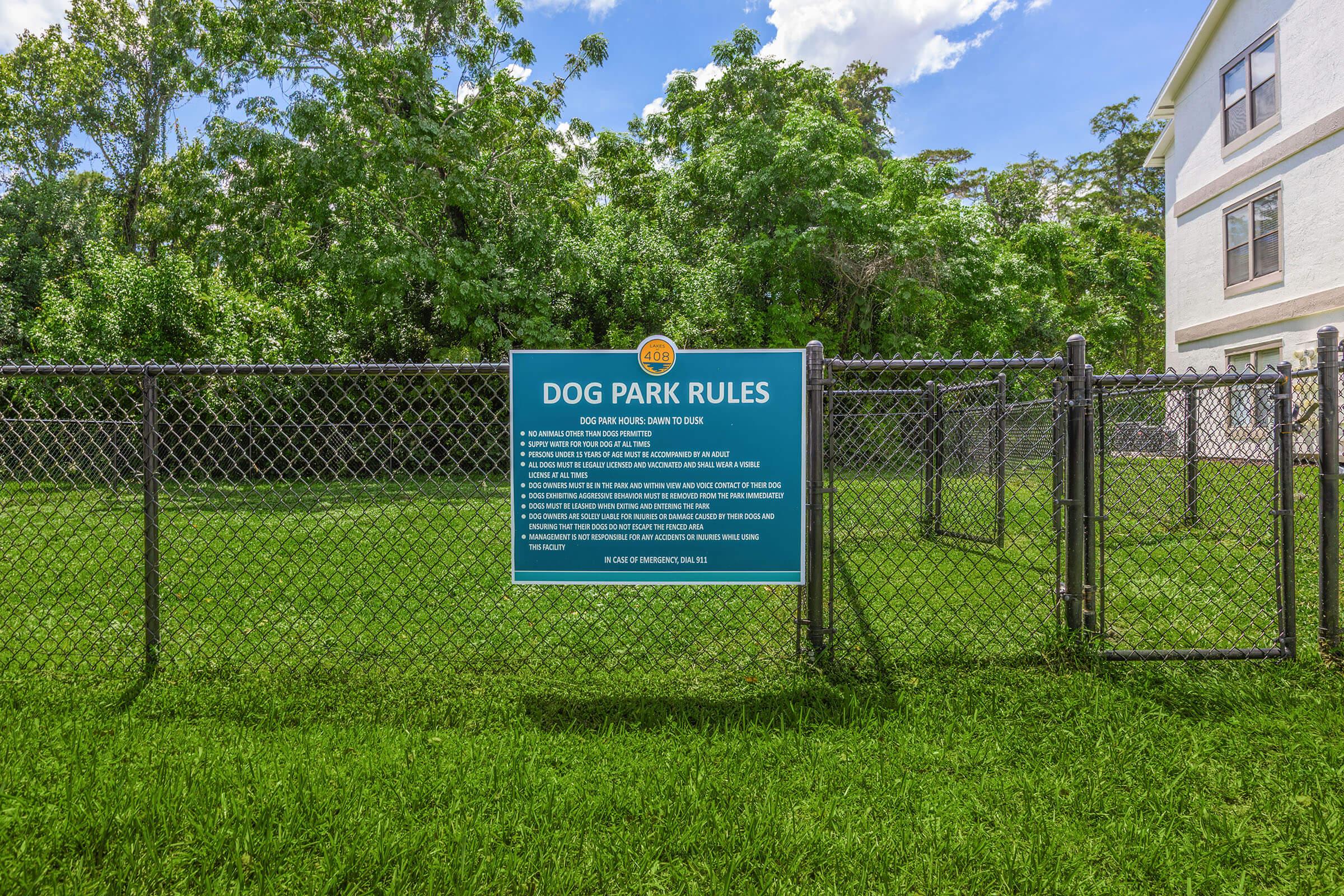 Sign displaying dog park rules on a fence. The rules include guidelines for dog owners regarding pet behavior, cleanliness, and safety. The background features a well-maintained grassy area with trees, and an adjacent building can be seen. Bright blue and yellow colors are used for the sign.