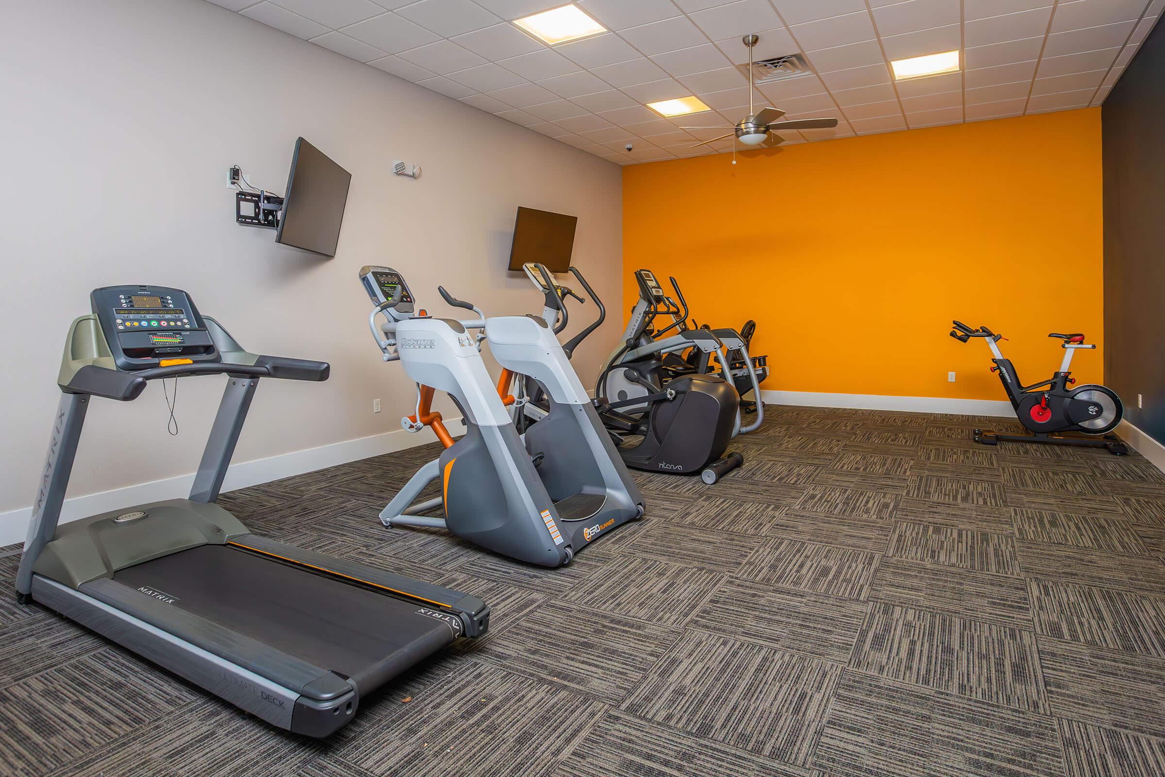 A modern fitness room featuring a treadmill, elliptical machines, and a stationary bike. The walls are painted in orange and gray tones, with flat-screen TVs mounted on the wall. The space has a clean, contemporary design with carpet flooring, providing a well-lit environment for exercise.