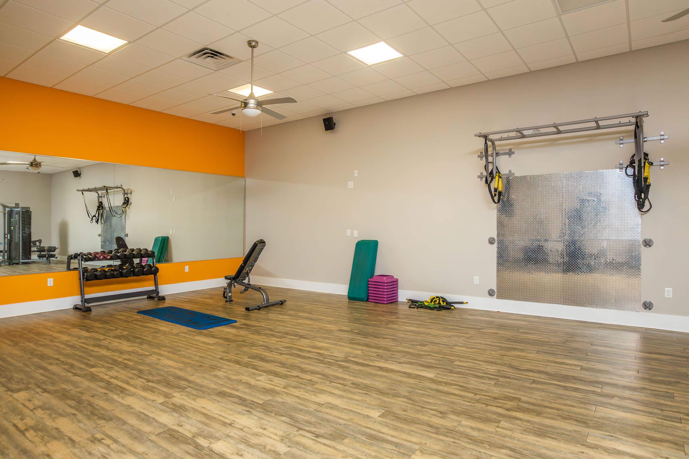 A well-lit fitness room featuring weights on a rack, a bench for exercises, a yoga mat, and colorful exercise equipment. There is a large mirror on one wall and a wall-mounted suspension training system, all set on a wooden floor with an orange accent wall.